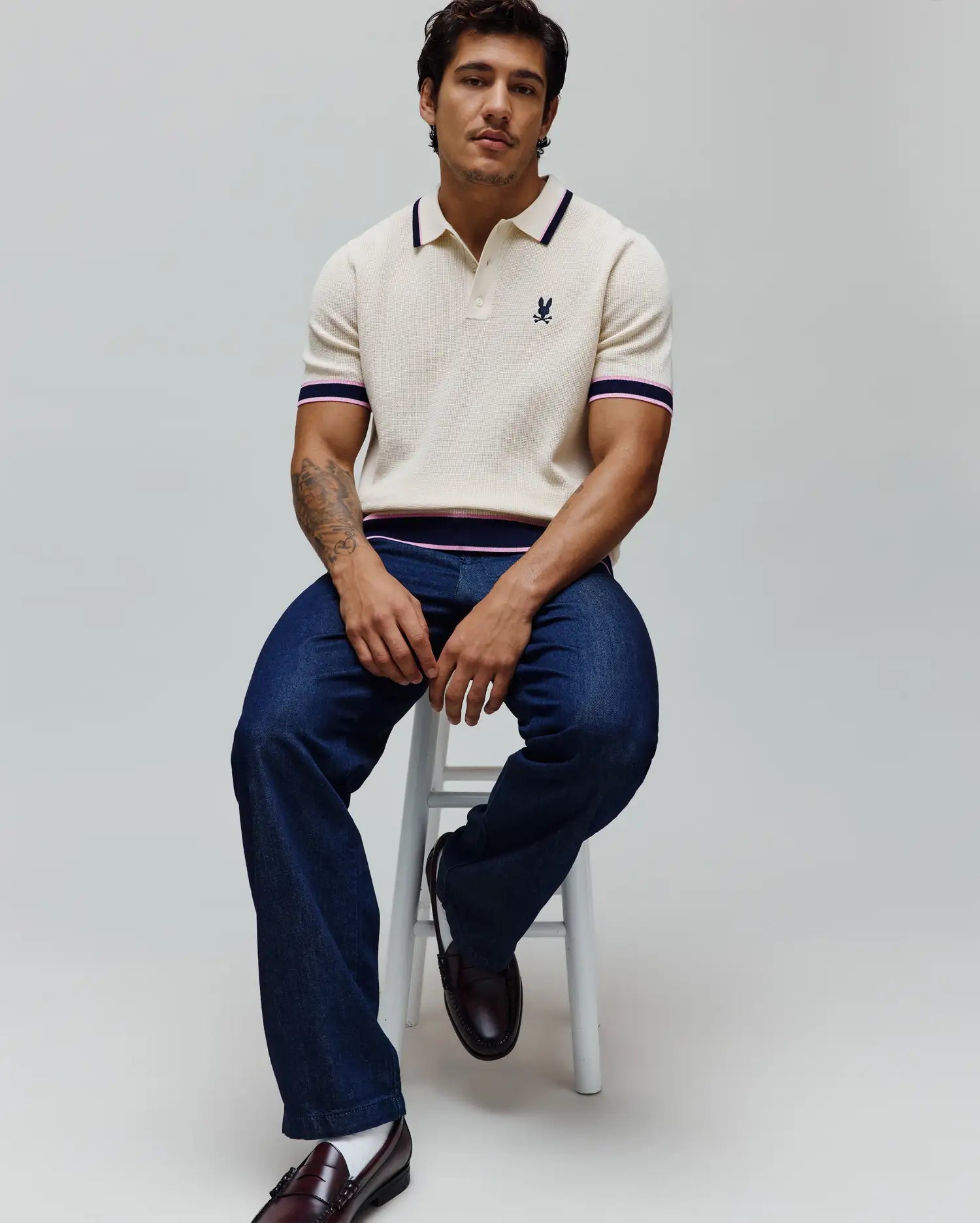 Mens Dax Knit Polo in cream with a classic fit, featuring a fine knit texture, navy and pink striped collar, sleeve cuffs, and hem, and a small embroidered Psycho Bunny skull logo on the left chest.