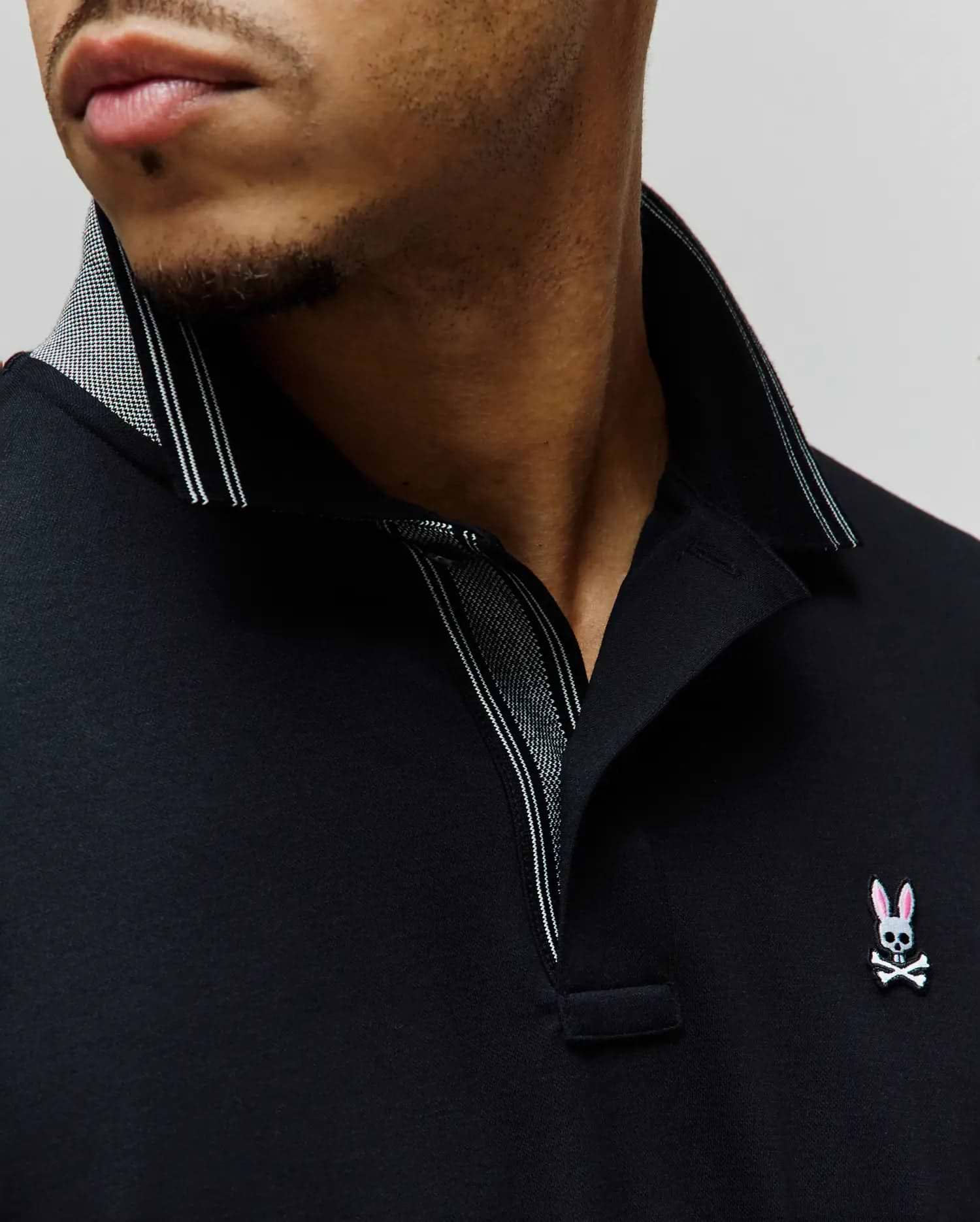 Mens Kenyon Lightweight Pique Polo in black, featuring a classic fit, textured pique fabric, and a small embroidered Psycho Bunny skull logo with pink ears on the left chest. The collar has a subtle houndstooth pattern and white stripe accents.