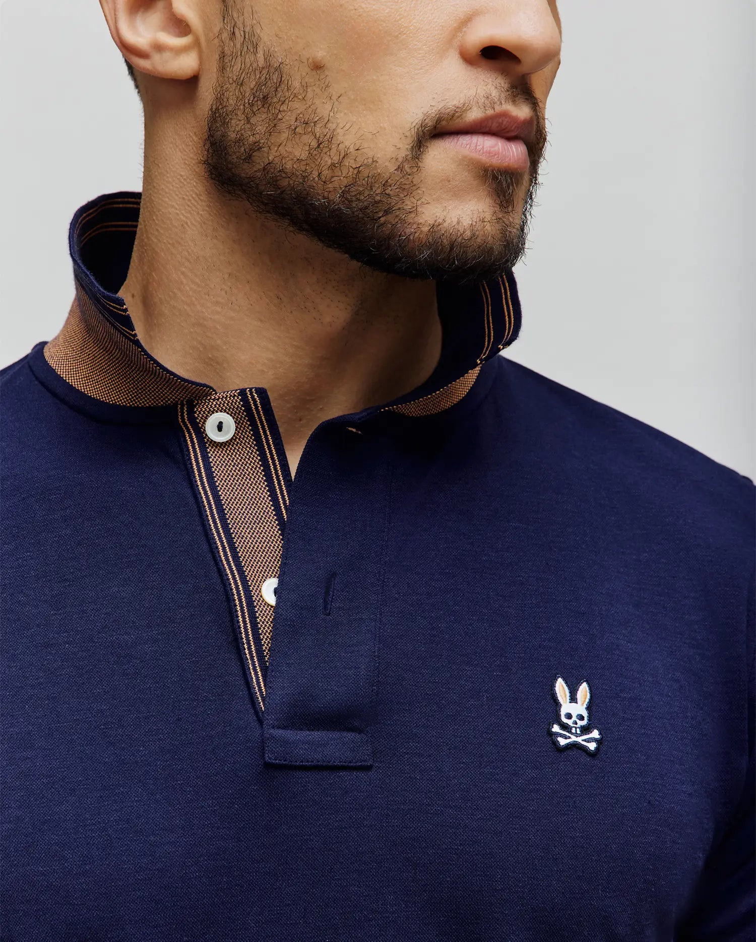 Mens Kenyon Lightweight Pique Polo in navy, featuring a classic fit, pique texture, and a small embroidered white and black Psycho Bunny skull logo on the left chest. The collar and placket have a contrasting brown and tan striped lining.