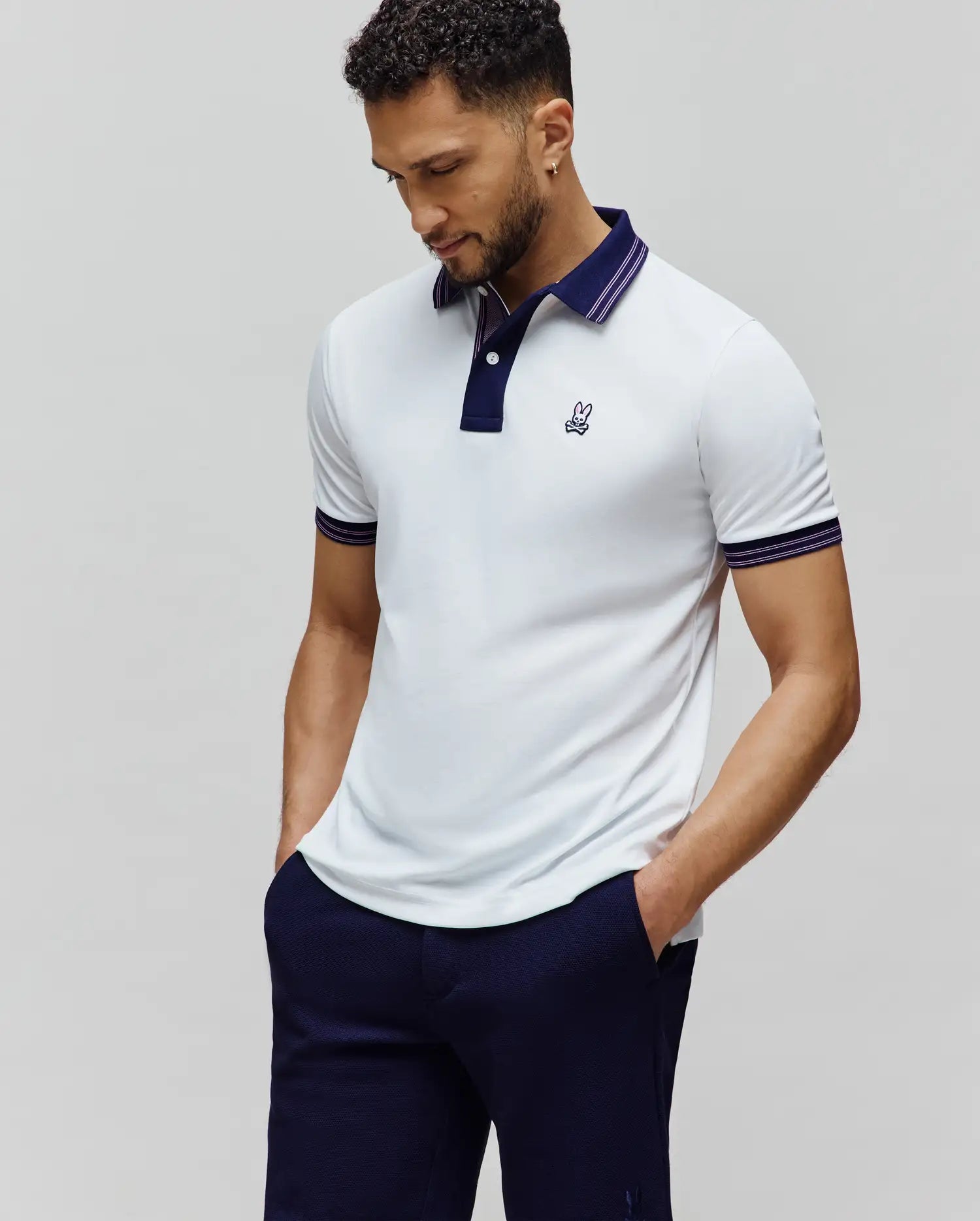 Mens Kenyon Lightweight Pique Polo, white pique with a classic fit, navy collar and placket, purple-striped cuffs, and an embroidered Psycho Bunny skull logo on the left chest.