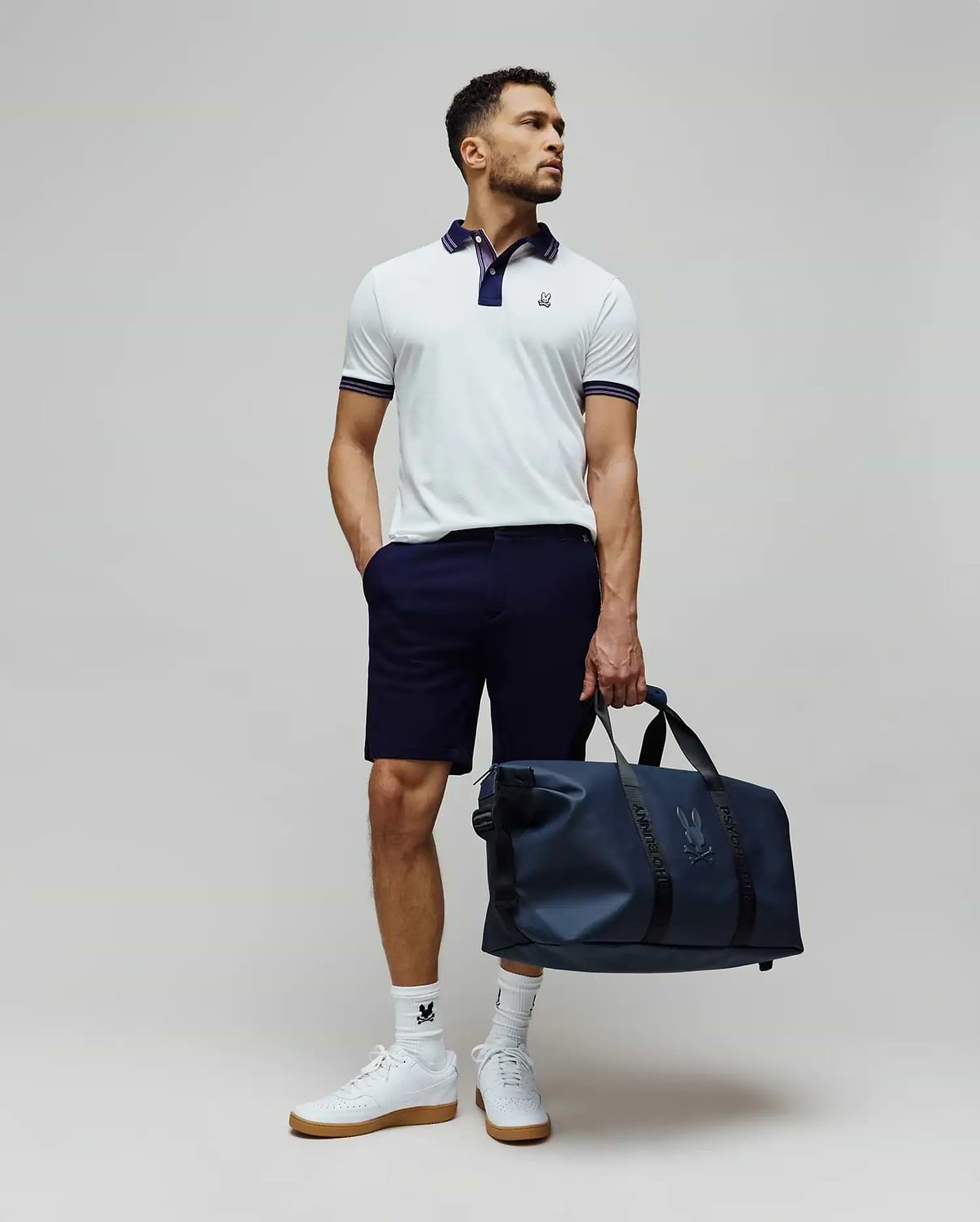Mens Kenyon Lightweight Pique Polo in white, featuring a navy collar, striped cuffs, and an embroidered Psycho Bunny logo on the chest. Styled with navy shorts, logo socks, and a matching navy duffle bag.