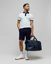 Mens Kenyon Lightweight Pique Polo in white, featuring a navy collar, striped cuffs, and an embroidered Psycho Bunny logo on the chest. Styled with navy shorts, logo socks, and a matching navy duffle bag.