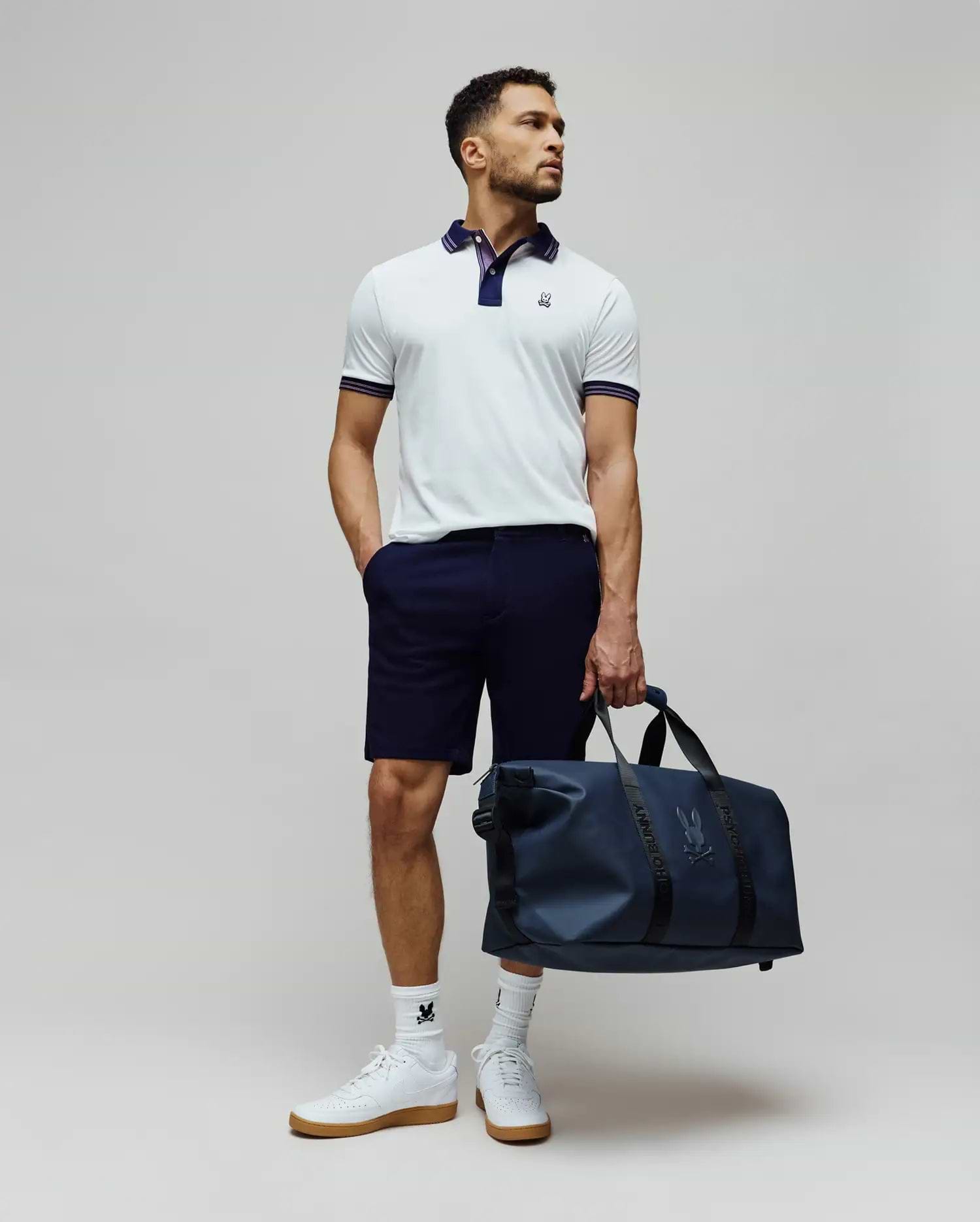 Mens Kenyon Lightweight Pique Polo in white, featuring a navy collar, striped cuffs, and an embroidered Psycho Bunny logo on the chest. Styled with navy shorts, logo socks, and a matching navy duffle bag.