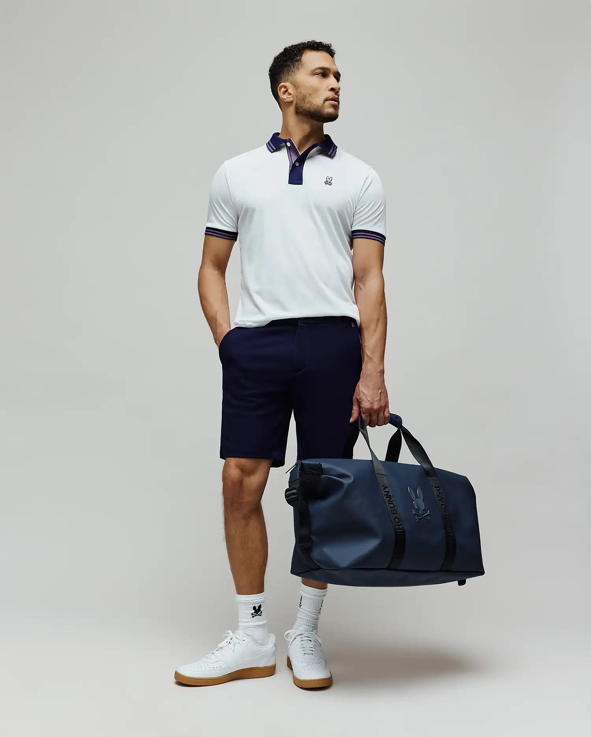 Mens Kenyon Lightweight Pique Polo in white, featuring a navy collar, striped cuffs, and an embroidered Psycho Bunny logo on the chest. Styled with navy shorts, logo socks, and a matching navy duffle bag.