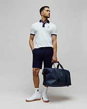Mens Kenyon Lightweight Pique Polo in white, featuring a navy collar, striped cuffs, and an embroidered Psycho Bunny logo on the chest. Styled with navy shorts, logo socks, and a matching navy duffle bag.