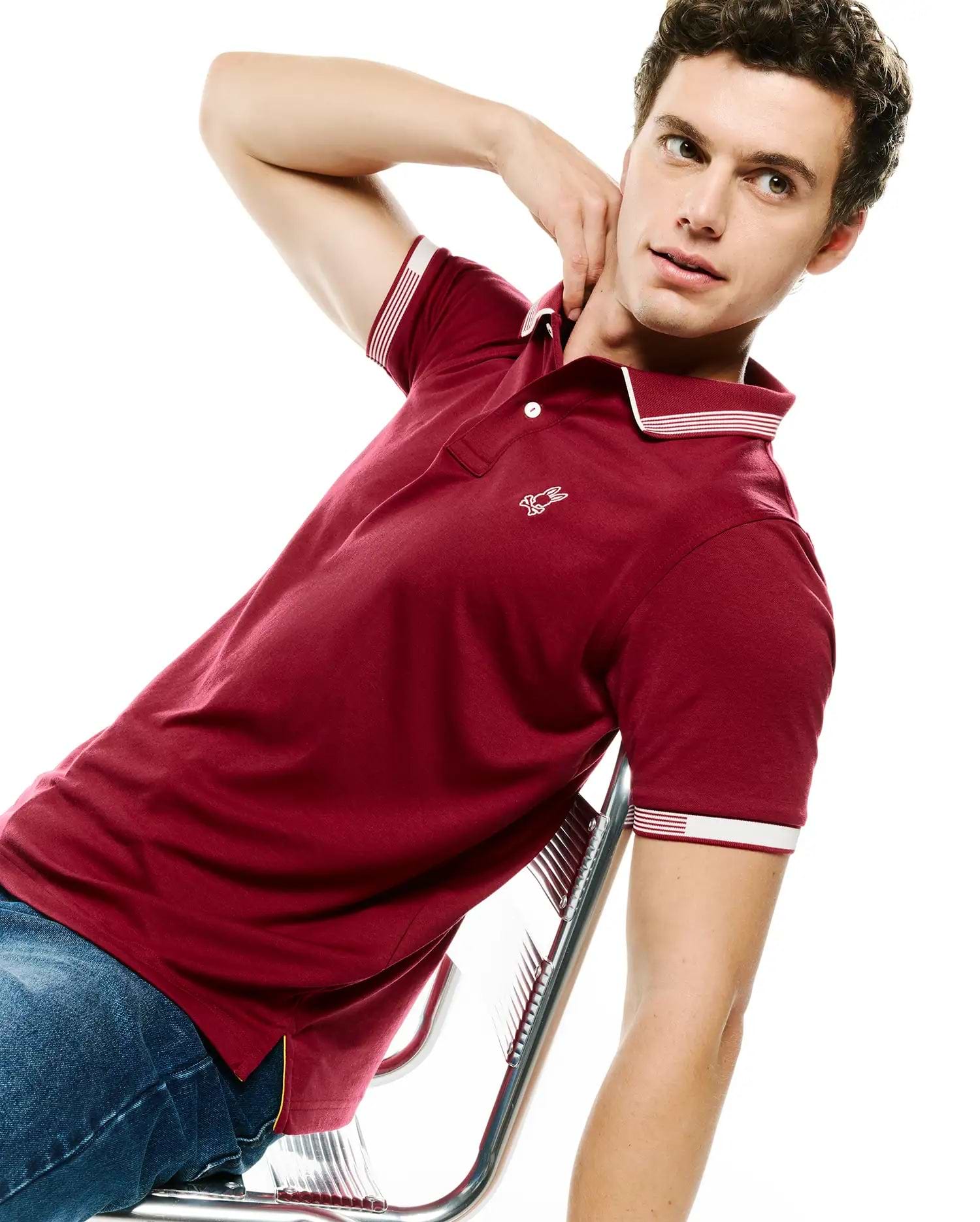 Mens Timmy Interlock Polo, a classic fit maroon interlock polo with a small embroidered white Psycho Bunny skull logo on the left chest, striped white and red ribbed collar and cuffs, and a two-button placket.