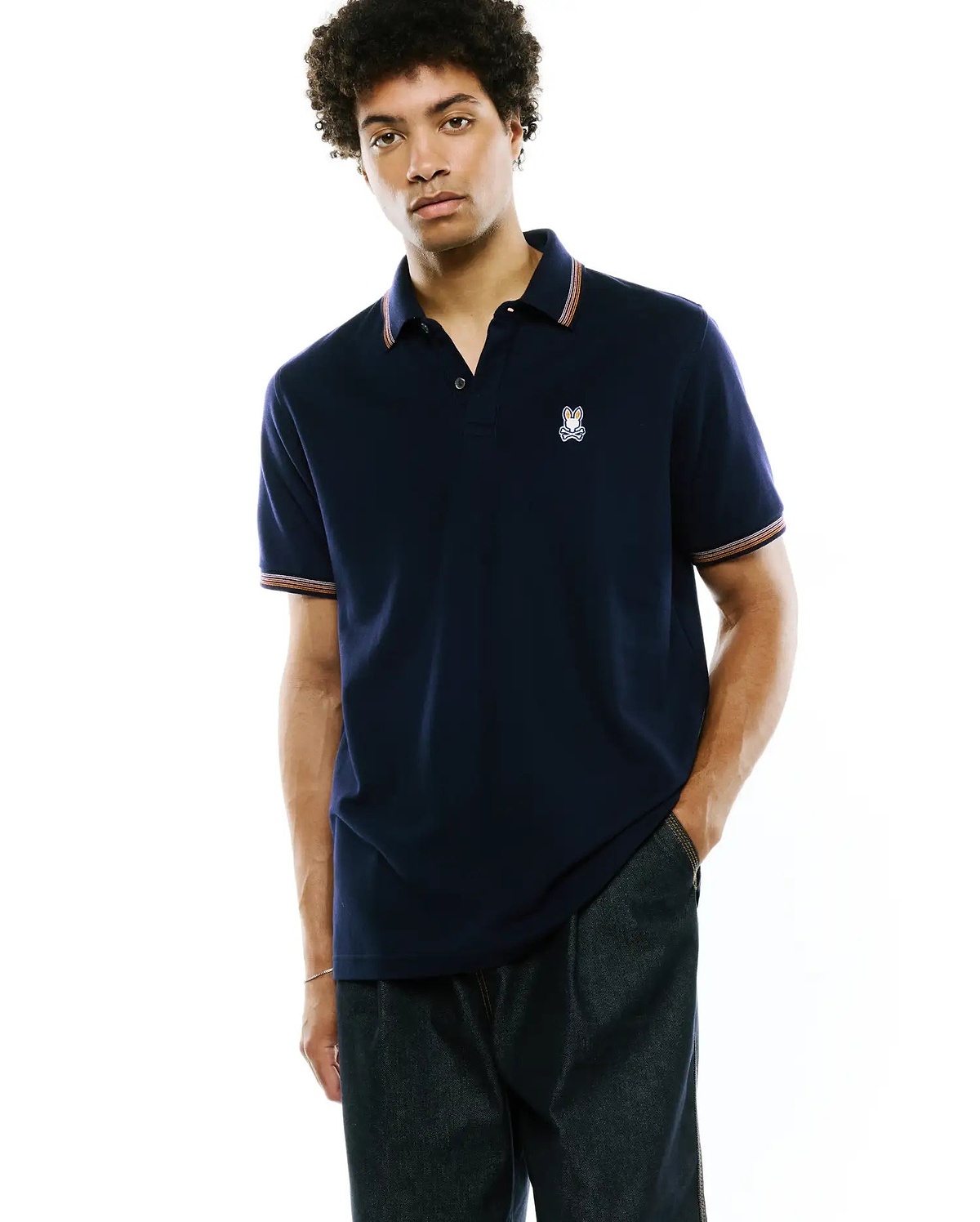 Mens Jaydon Pique Polo, navy classic fit pique knit with a small embroidered Psycho Bunny skull logo on the left chest and striped collar and sleeve cuffs in orange, white, and blue.
