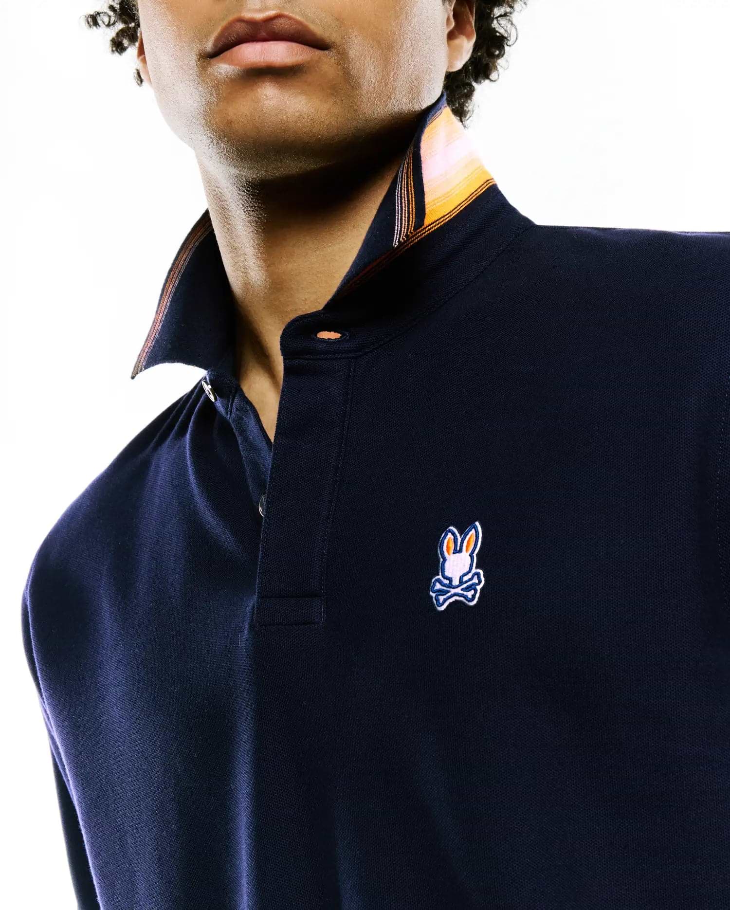 Mens Jaydon Pique Polo. Navy blue classic fit pique polo with a small embroidered Psycho Bunny skull logo on the left chest. Features a striped inner collar in orange and yellow, and orange buttons.
