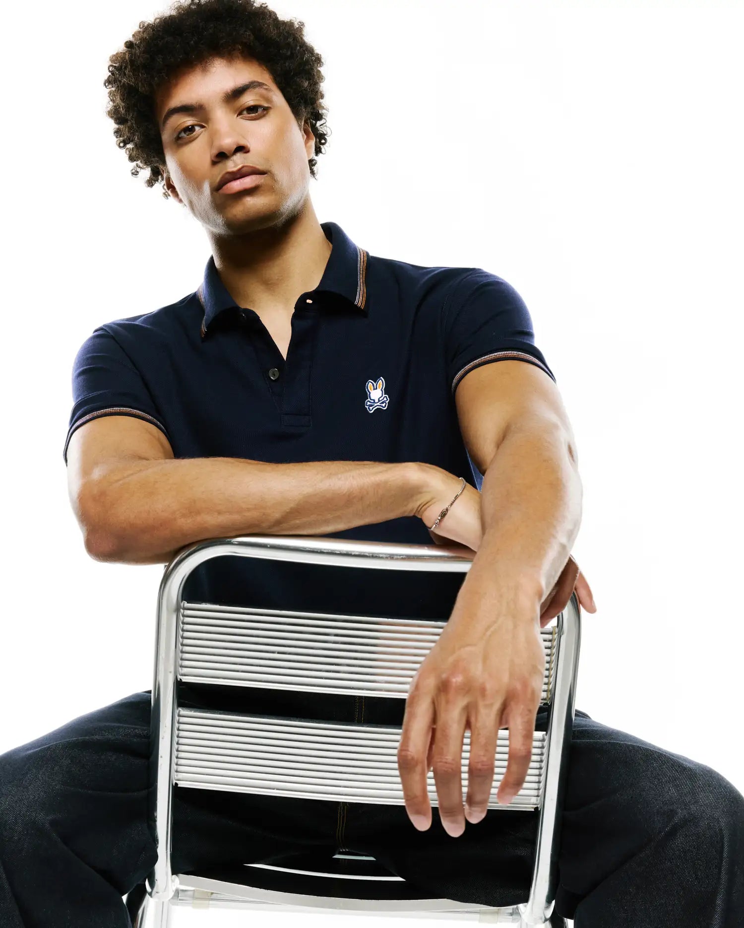 Mens Jaydon Pique Polo, navy classic fit pique polo with orange and white striped trim on collar and cuffs, featuring a small embroidered Psycho Bunny skull logo on the left chest.