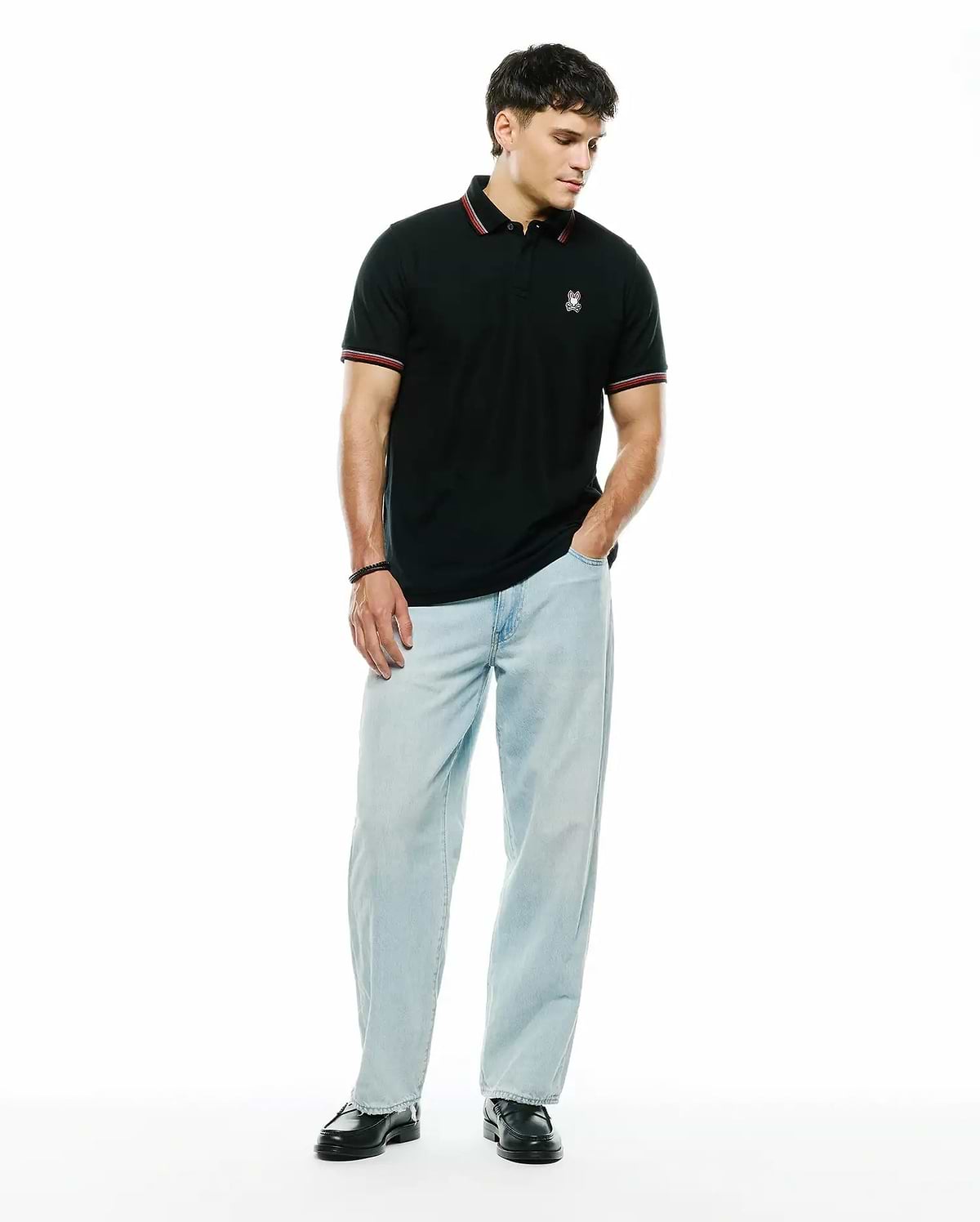 Mens Jaydon Pique Polo, a classic fit black pique polo with a small embroidered white and red Psycho Bunny skull logo on the left chest, featuring red and white striped collar and sleeve cuffs.