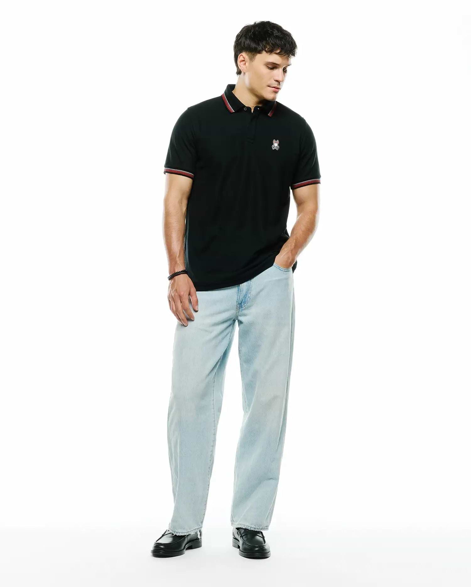 Mens Jaydon Pique Polo, a classic fit black pique polo with a small embroidered white and red Psycho Bunny skull logo on the left chest, featuring red and white striped collar and sleeve cuffs.