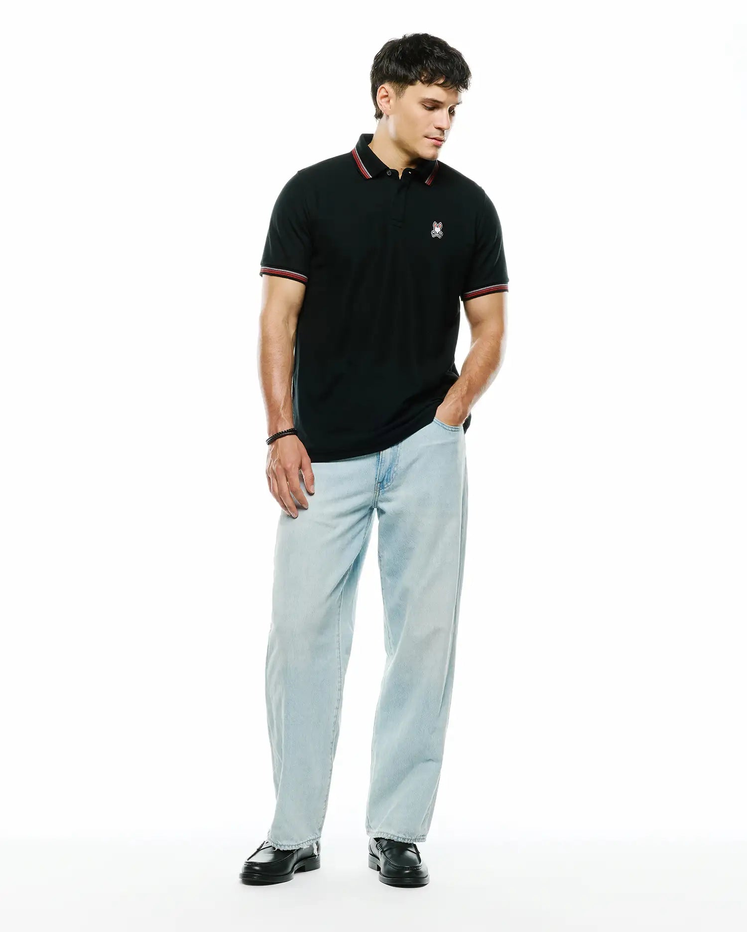 Mens Jaydon Pique Polo, a classic fit black pique polo with a small embroidered white and red Psycho Bunny skull logo on the left chest, featuring red and white striped collar and sleeve cuffs.