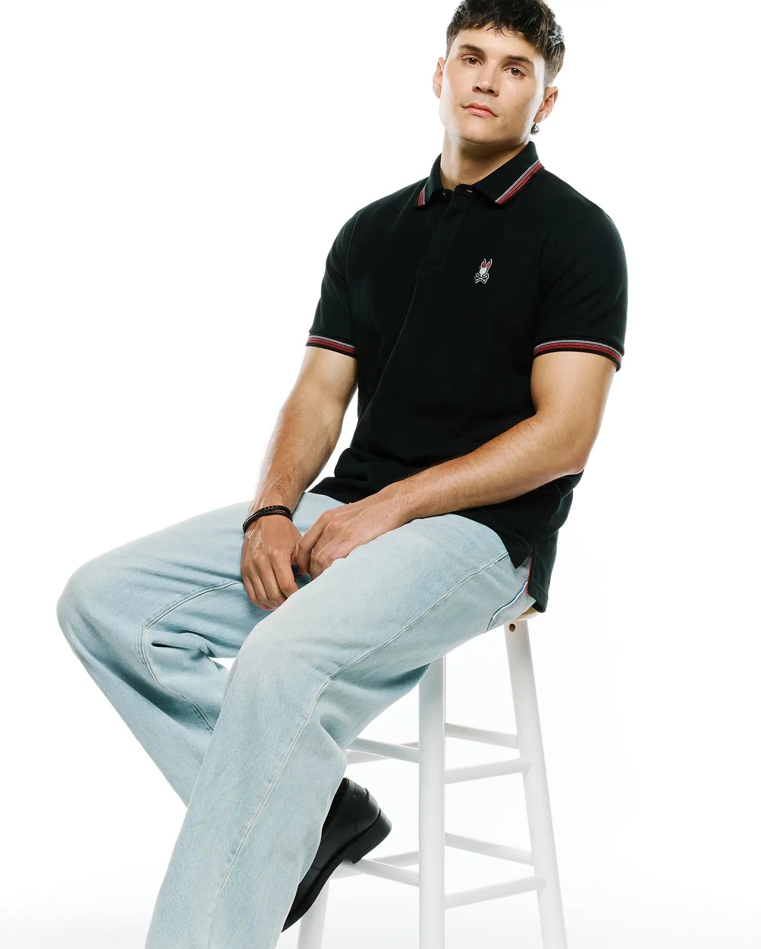 Mens Jaydon Pique Polo in black, featuring a classic fit, textured pique fabric, red and white striped collar and cuffs, and a small embroidered Psycho Bunny skull logo on the left chest.