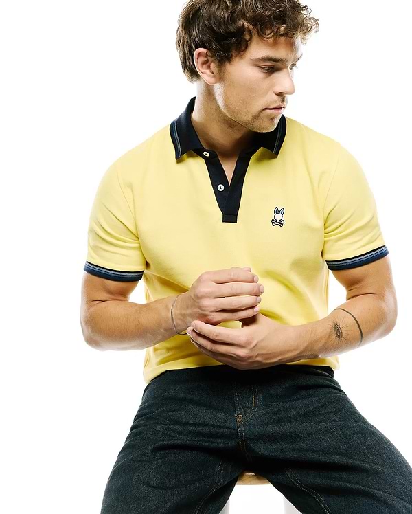 Mens Jaydon Pique Polo, classic fit, yellow pique with navy collar, placket, and striped cuffs. Features a small embroidered Psycho Bunny skull logo with blue ears on the left chest.