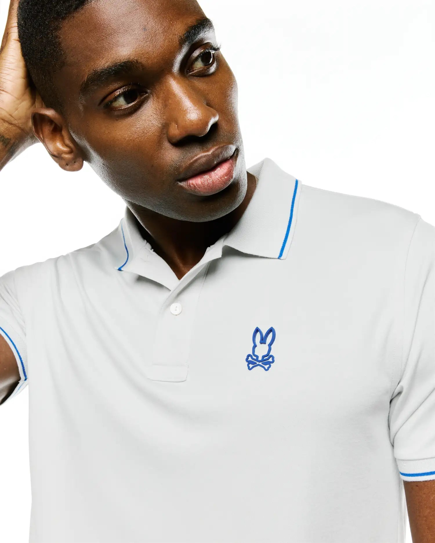 Men's Joji Performance Polo, light grey pique knit with a classic fit, features a two-button placket, blue tipping on the collar and sleeve cuffs, and an embroidered blue Psycho Bunny skull logo on the left chest.
