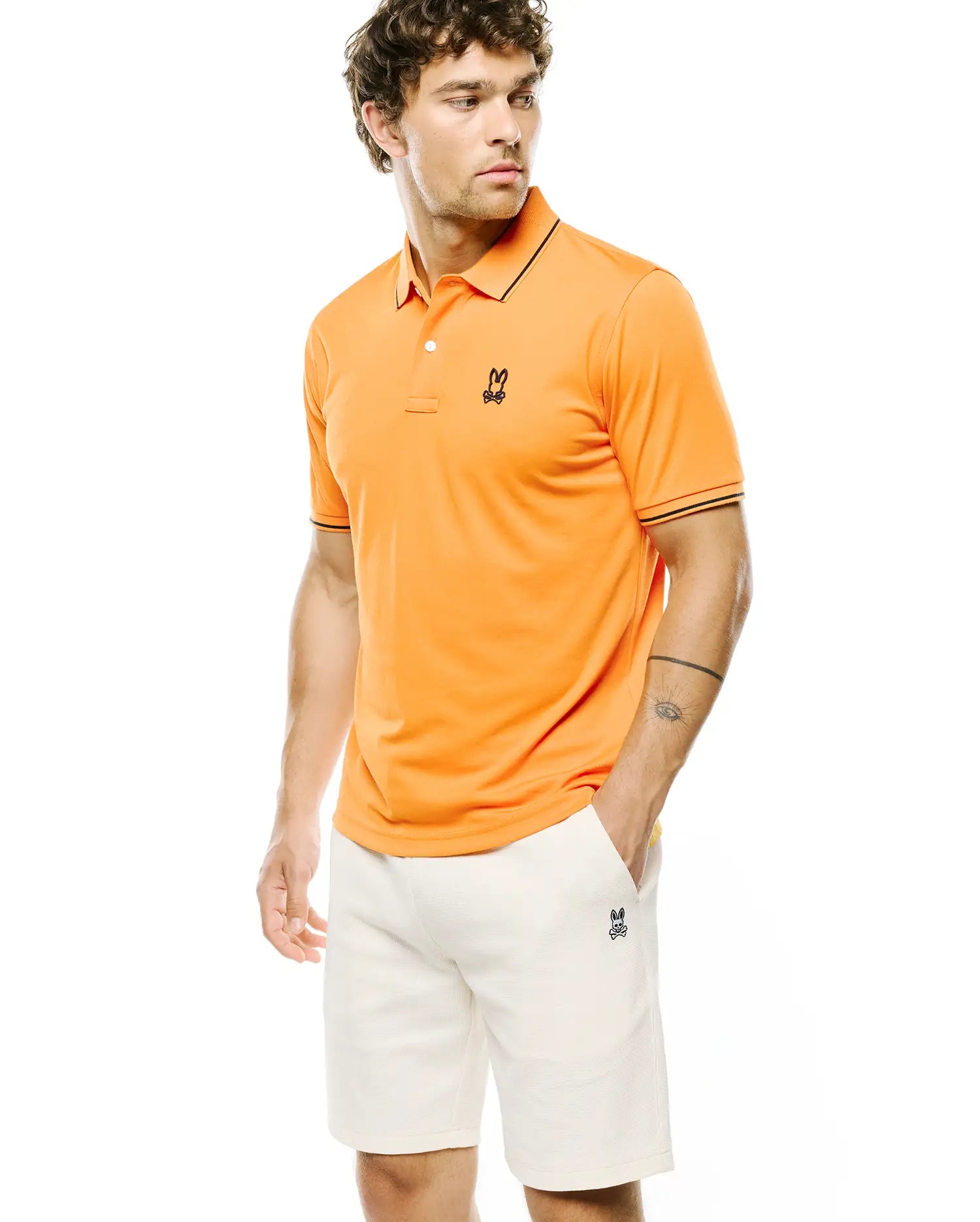 Bright Orange Mens Joji Performance Polo with black tipping on collar and cuffs, featuring a small embroidered black Psycho Bunny logo on the left chest, paired with off-white textured shorts with a matching embroidered logo.