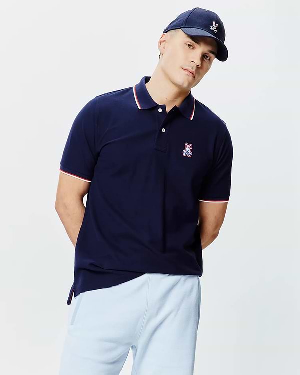 Navy blue Mens John Pique Polo, classic fit, with white two-button placket, ribbed collar and cuffs featuring red and white tipping, and an embroidered Psycho Bunny skull logo on the left chest.