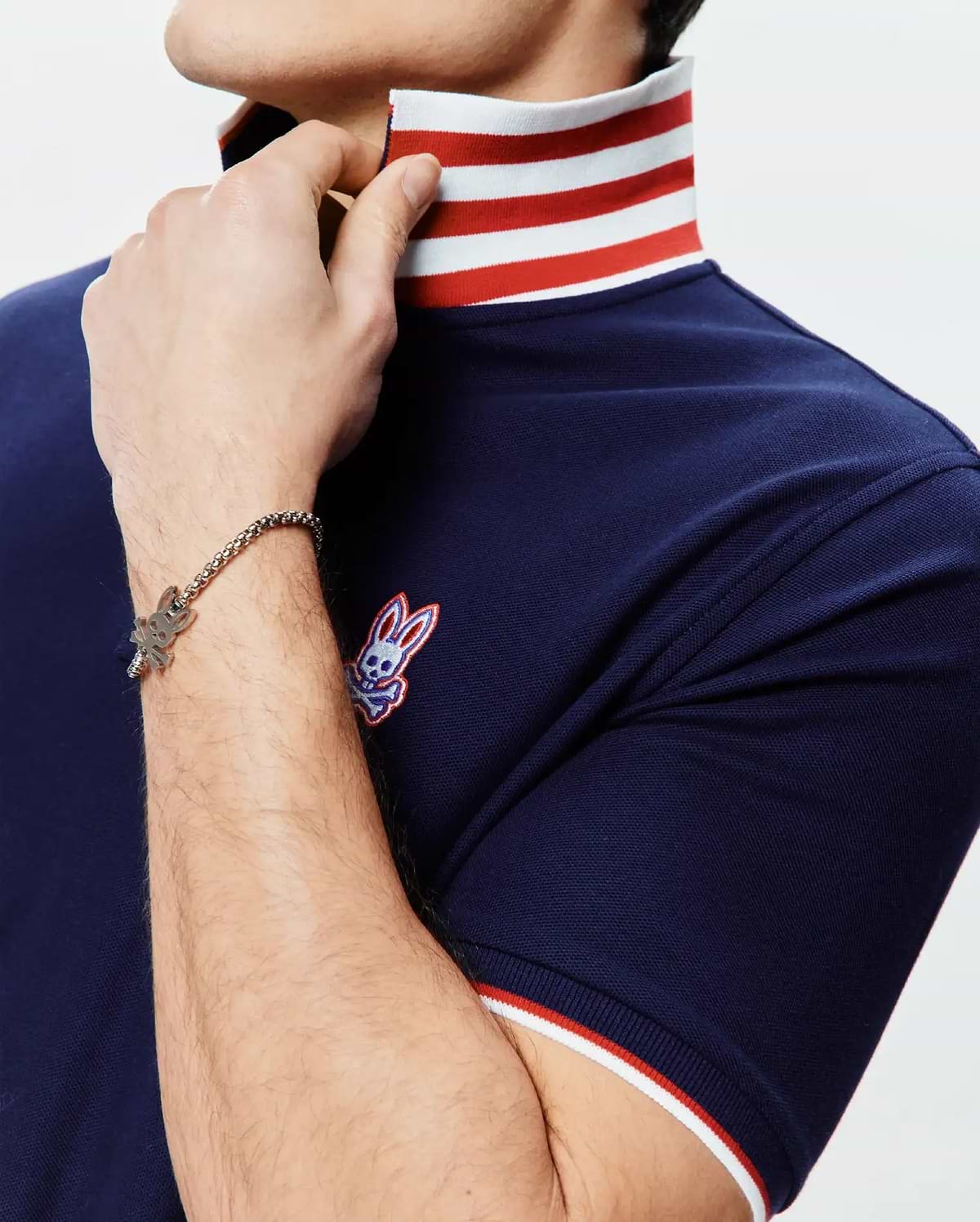 Mens John Pique Polo, a navy blue pique polo with a classic fit, featuring a white and red striped collar and sleeve cuffs, and an embroidered Psycho Bunny skull logo on the left chest.