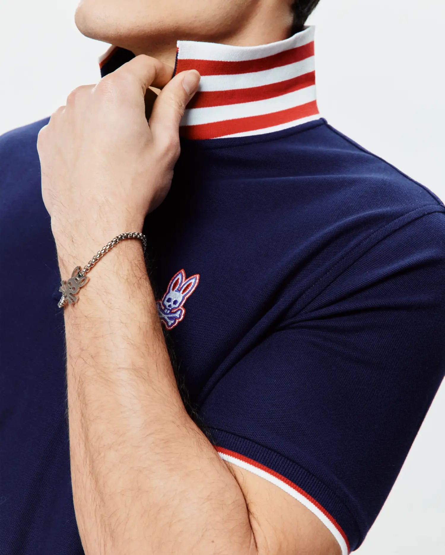 Mens John Pique Polo, a navy blue pique polo with a classic fit, featuring a white and red striped collar and sleeve cuffs, and an embroidered Psycho Bunny skull logo on the left chest.