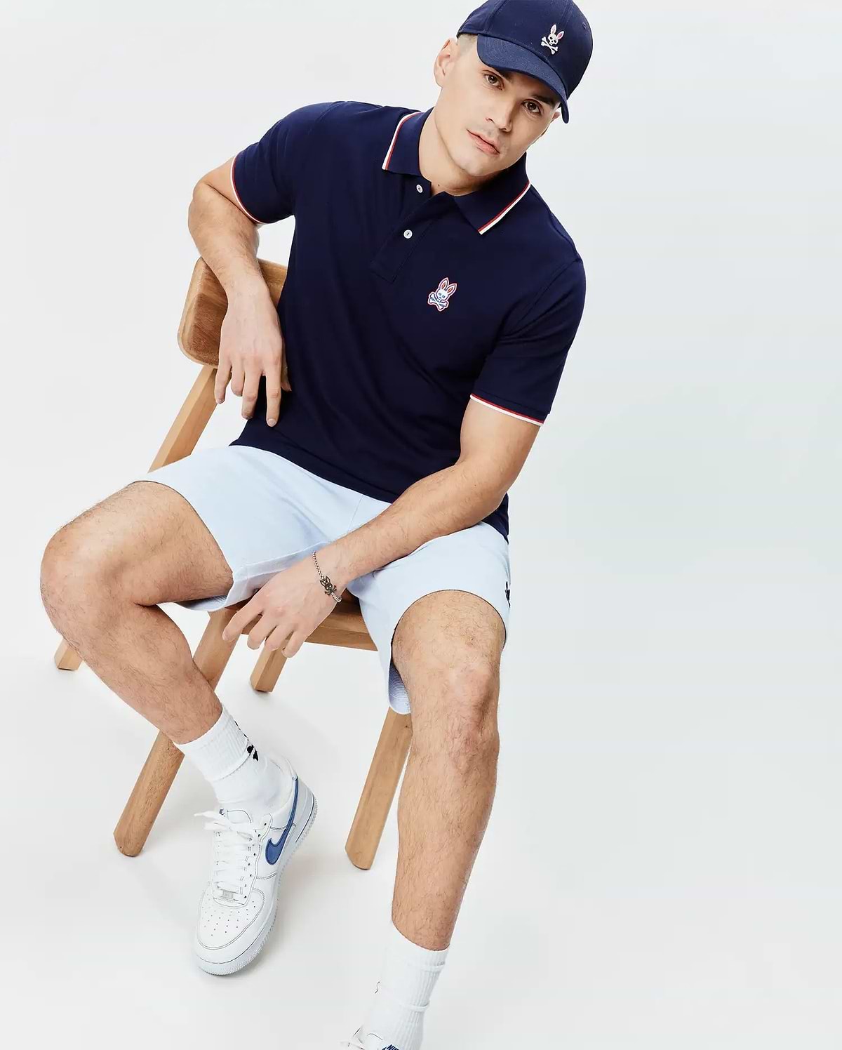 Mens John Pique Polo in navy blue with white and red tipping on collar and cuffs, featuring a small embroidered Psycho Bunny logo on the left chest. Paired with light blue shorts and a navy cap with an embroidered bunny skull.