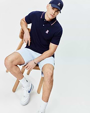 Mens John Pique Polo in navy blue with white and red tipping on collar and cuffs, featuring a small embroidered Psycho Bunny logo on the left chest. Paired with light blue shorts and a navy cap with an embroidered bunny skull.