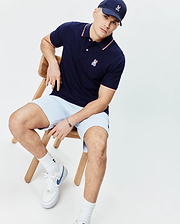 Mens John Pique Polo in navy blue with white and red tipping on collar and cuffs, featuring a small embroidered Psycho Bunny logo on the left chest. Paired with light blue shorts and a navy cap with an embroidered bunny skull.