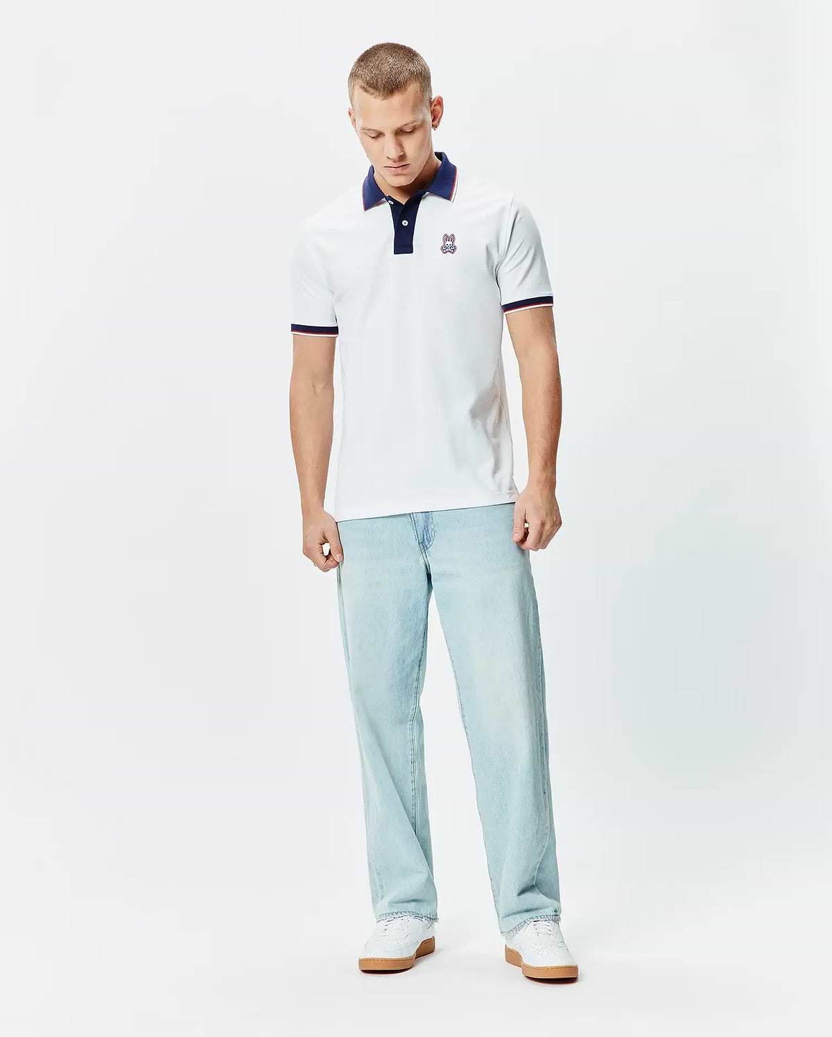 Mens John Pique Polo, a classic fit white pique polo with a navy collar, two-button placket, red and navy striped sleeve cuffs, and a small embroidered Psycho Bunny skull logo on the left chest.