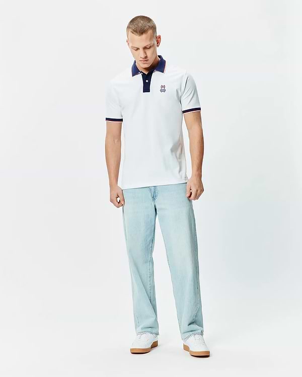 Mens John Pique Polo, a classic fit white pique polo with a navy collar, two-button placket, red and navy striped sleeve cuffs, and a small embroidered Psycho Bunny skull logo on the left chest.