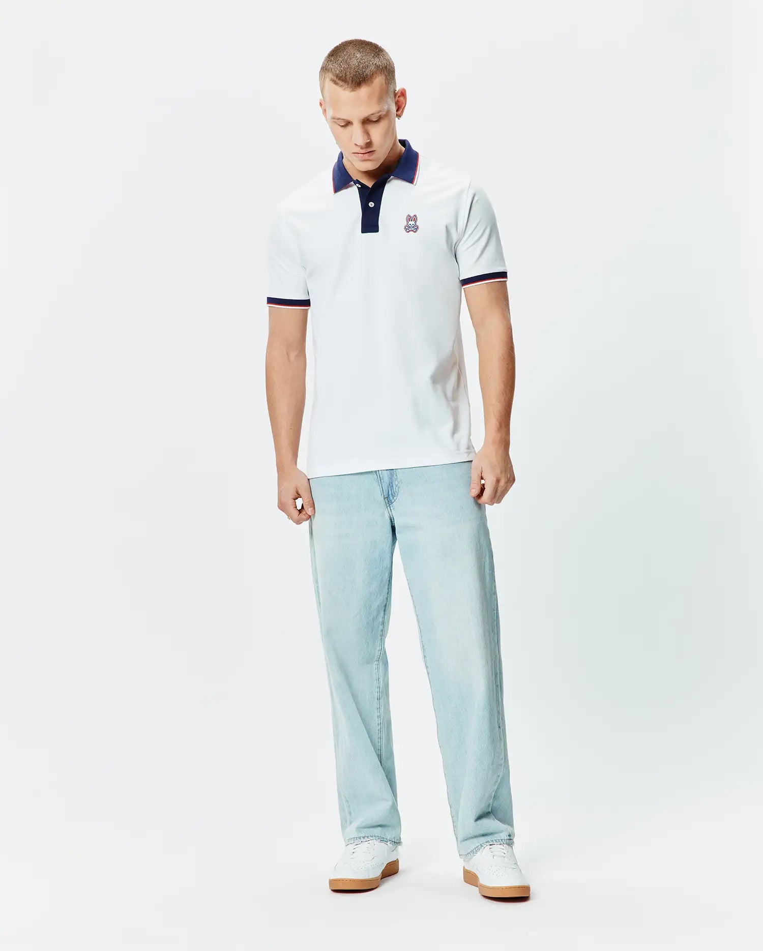 Mens John Pique Polo, a classic fit white pique polo with a navy collar, two-button placket, red and navy striped sleeve cuffs, and a small embroidered Psycho Bunny skull logo on the left chest.