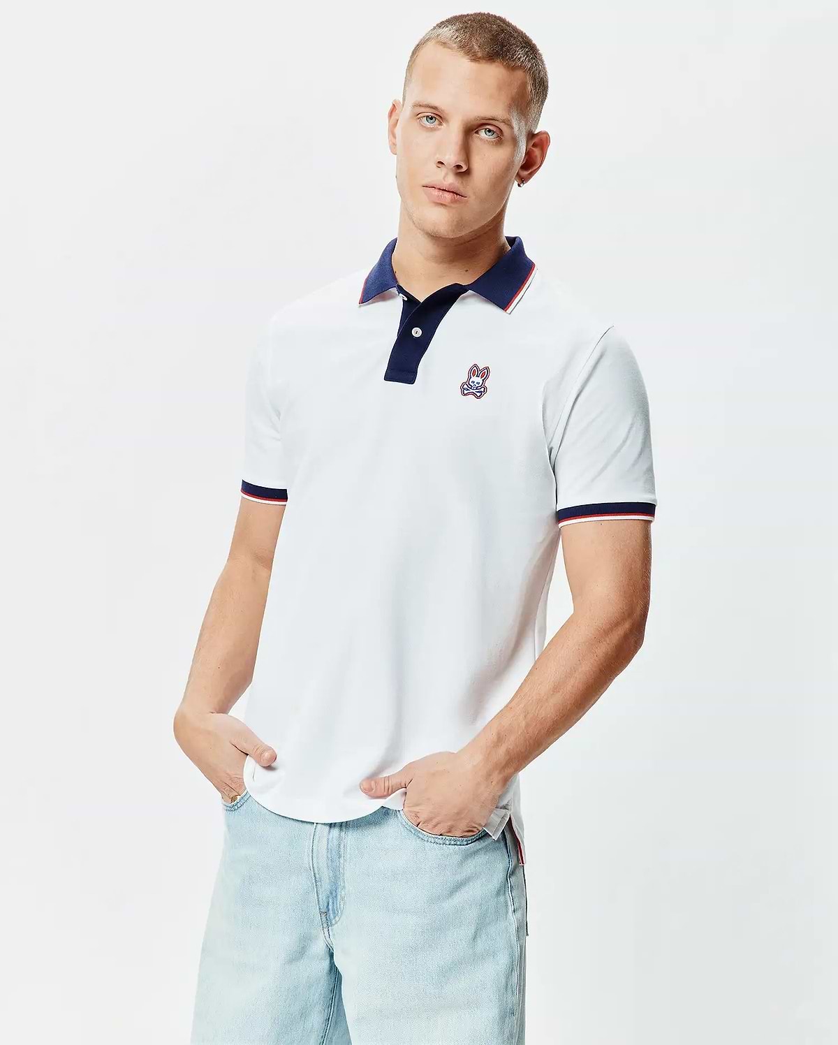 Mens John Pique Polo, a classic fit white pique polo shirt featuring a navy collar with red and white striped trim, matching striped cuffs, and a small embroidered Psycho Bunny skull logo on the left chest.