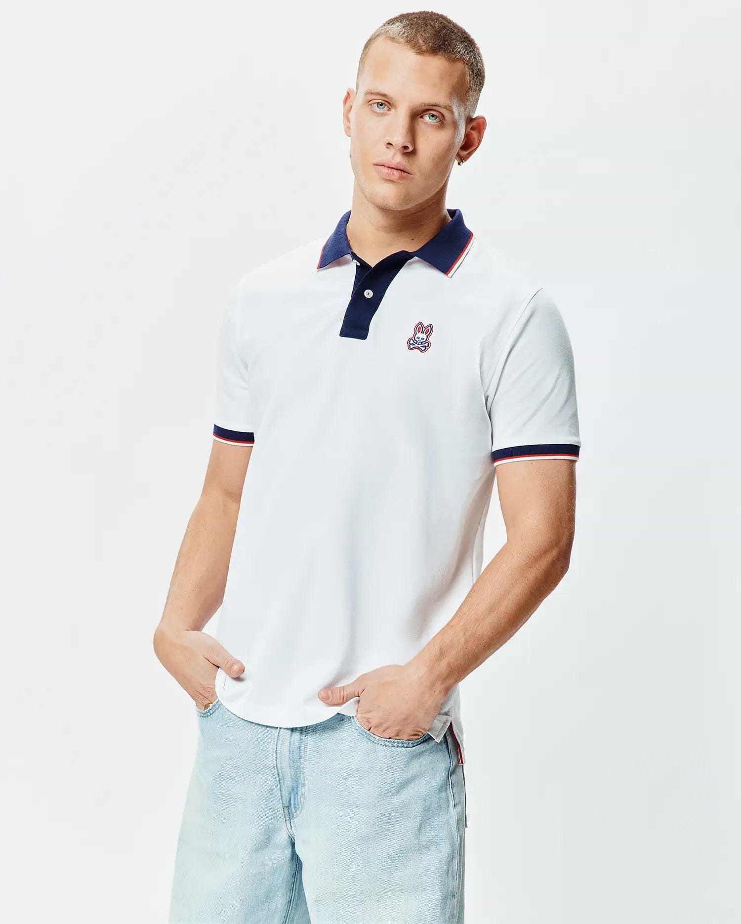 Mens John Pique Polo, a classic fit white pique polo shirt featuring a navy collar with red and white striped trim, matching striped cuffs, and a small embroidered Psycho Bunny skull logo on the left chest.
