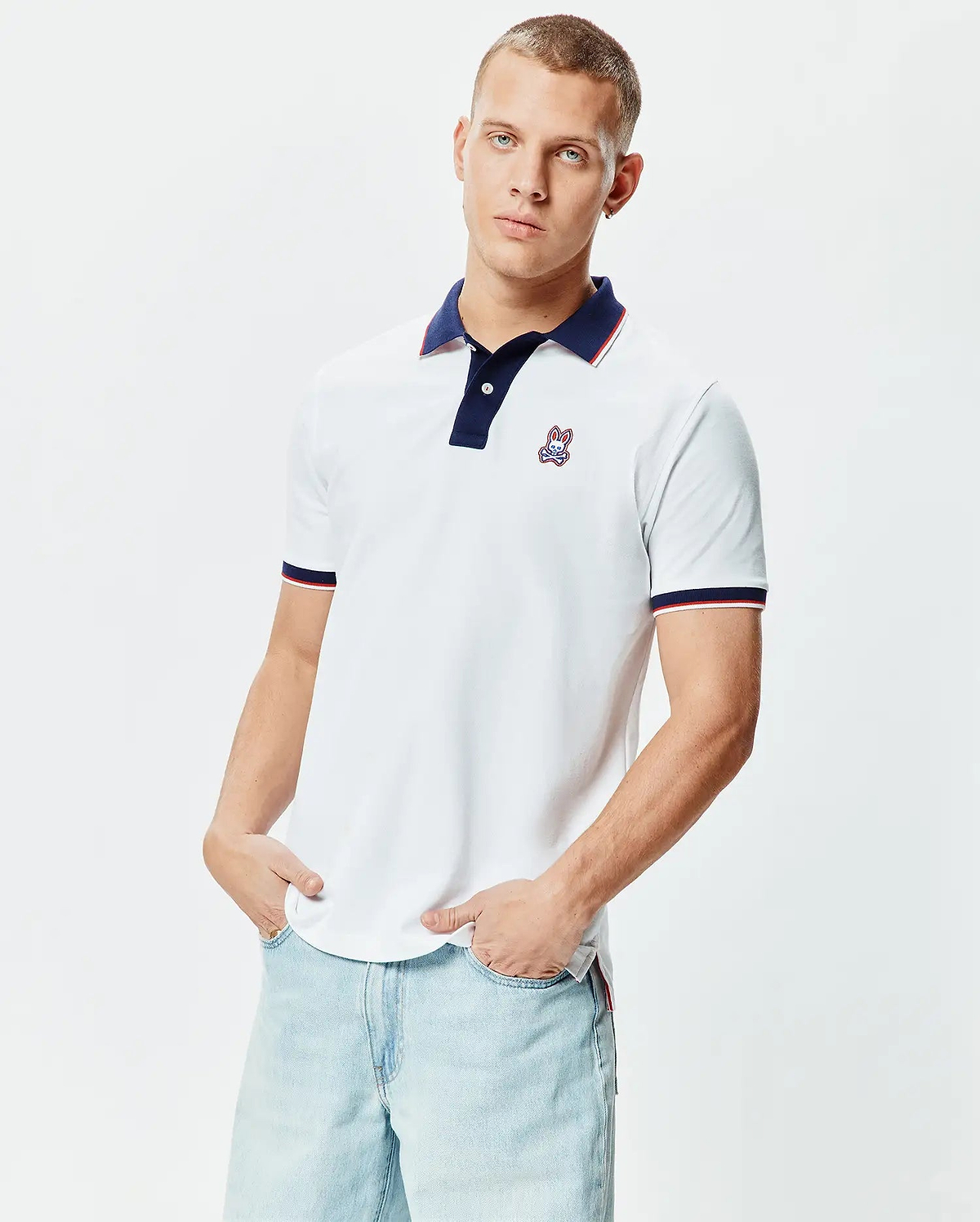 Mens John Pique Polo, a classic fit white pique polo shirt featuring a navy collar with red and white striped trim, matching striped cuffs, and a small embroidered Psycho Bunny skull logo on the left chest.