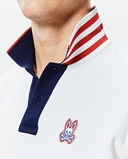 Mens John Pique Polo, a classic fit light blue pique polo with a navy placket, a bold red and white striped collar, and an embroidered Psycho Bunny skull logo on the left chest.
