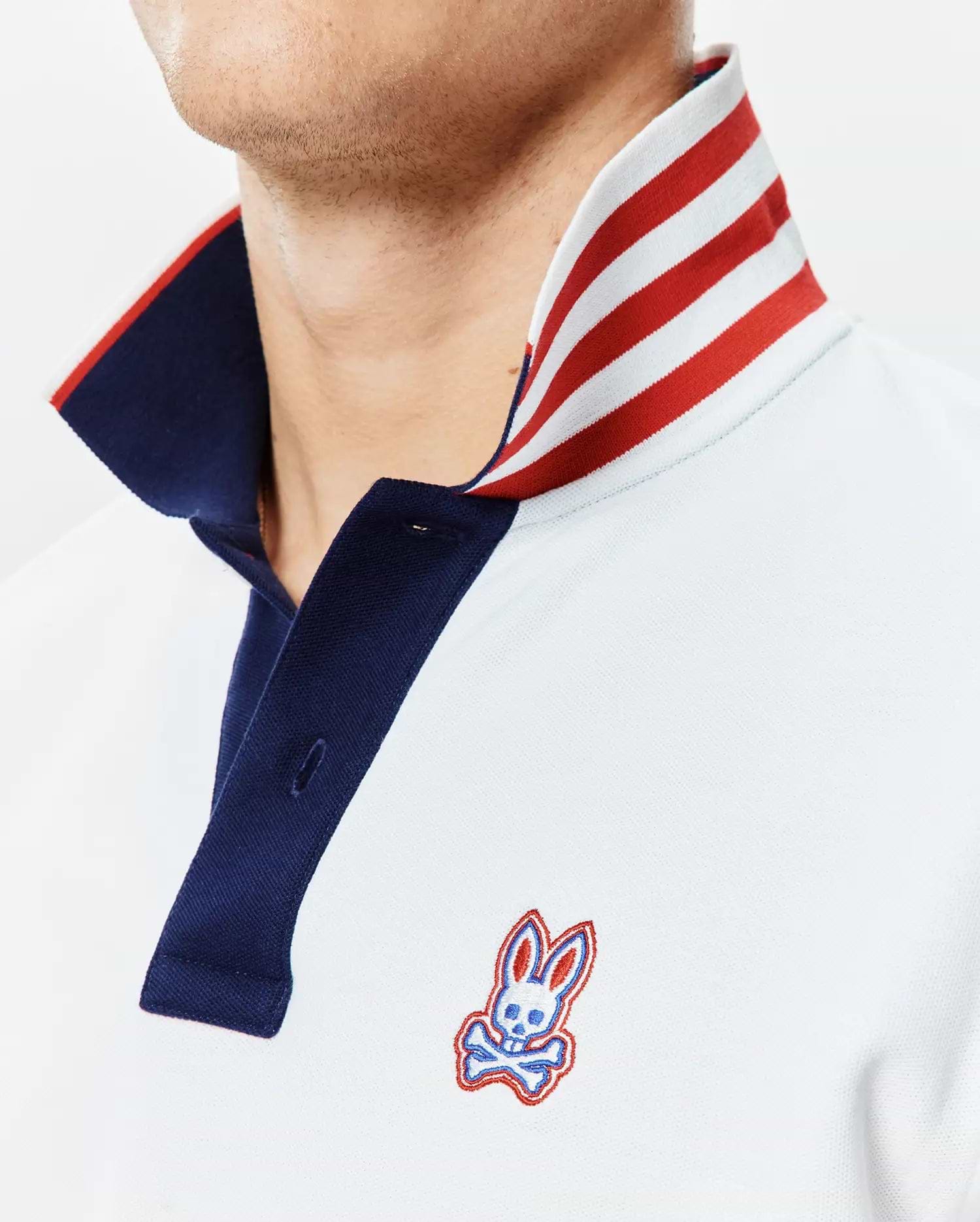 Mens John Pique Polo, a classic fit light blue pique polo with a navy placket, a bold red and white striped collar, and an embroidered Psycho Bunny skull logo on the left chest.