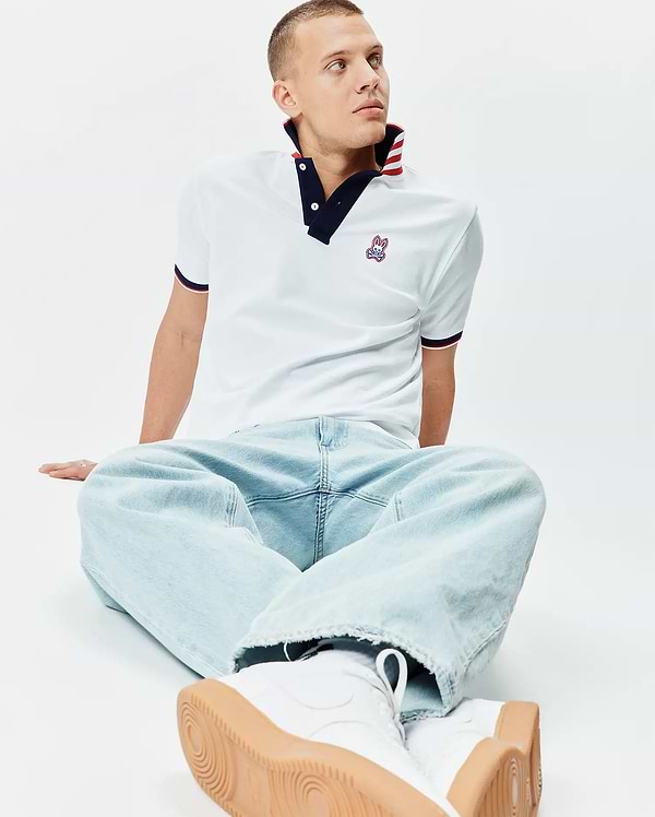 White Mens John Pique Polo, classic fit, with a navy collar featuring red and white stripes, matching striped cuffs, and an embroidered Psycho Bunny skull logo on the left chest.