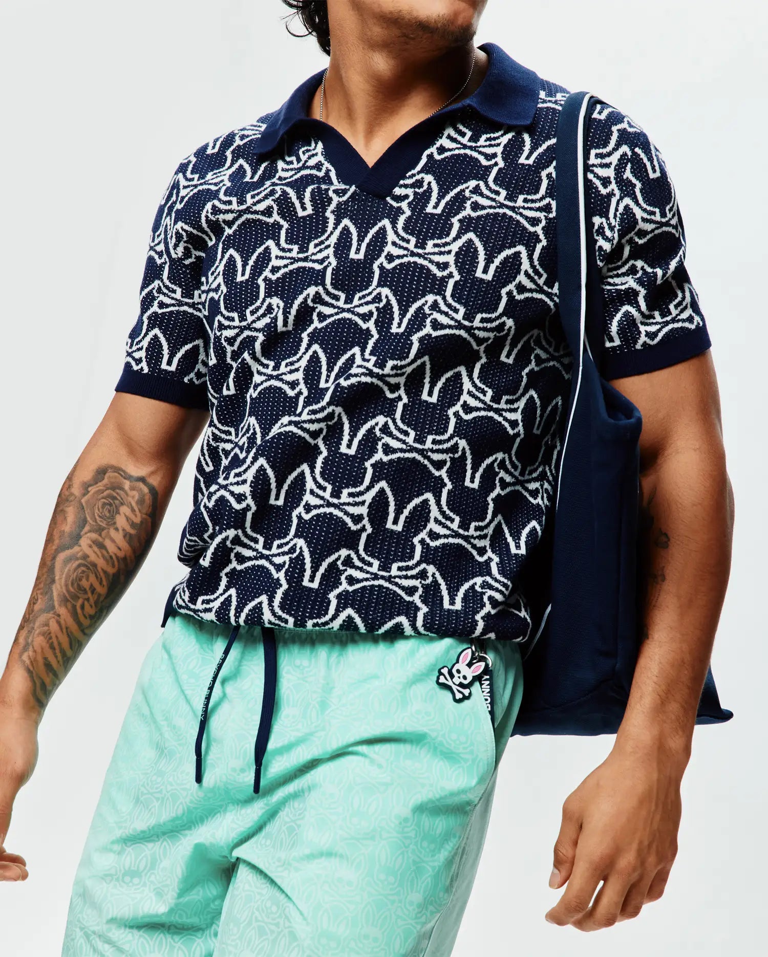Mens Sidney Oversized Knit Polo, a navy short-sleeve polo with an all-over white Psycho Bunny skull logo pattern, featuring a textured knit fabric, solid navy collar, and cuffs.
