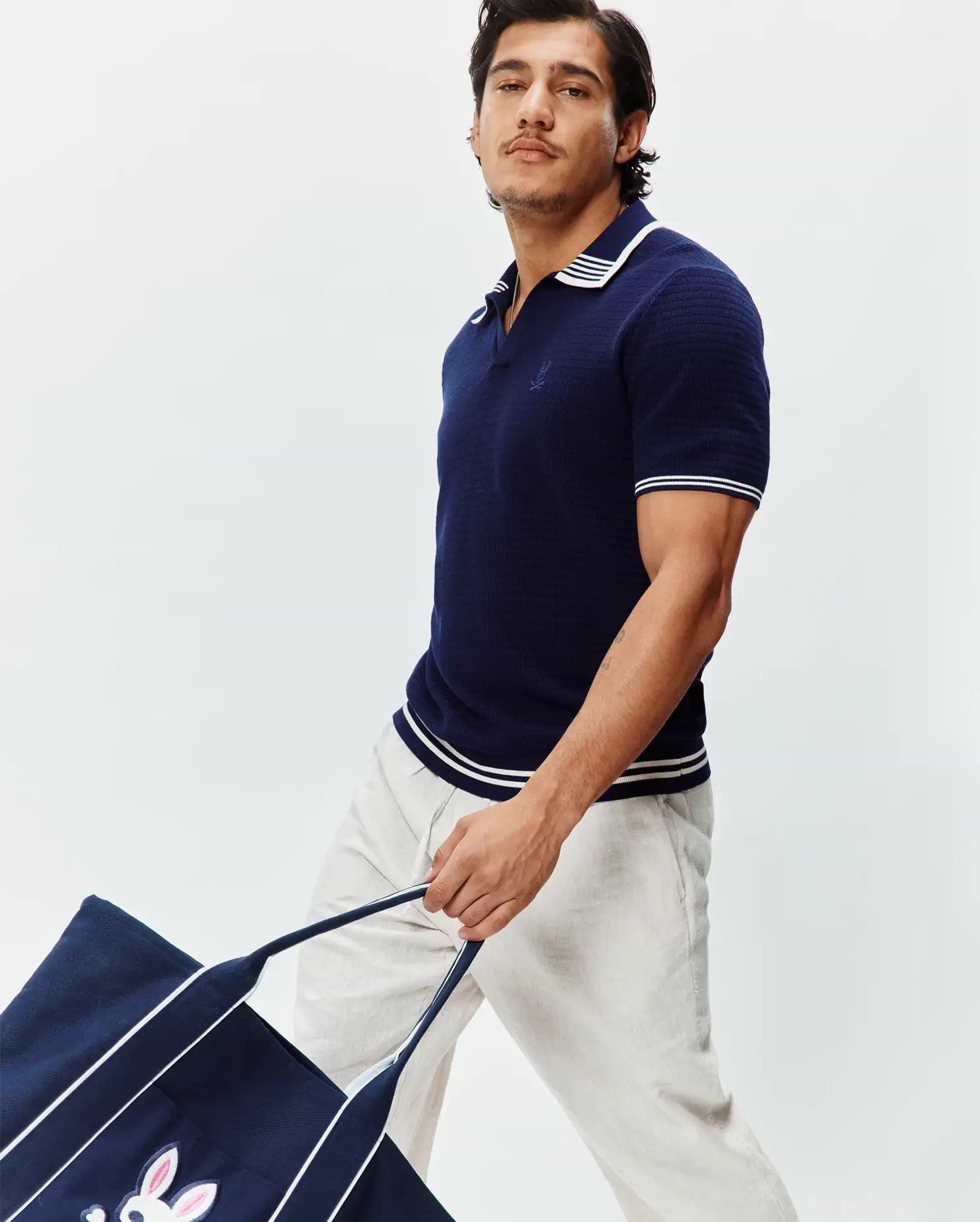 Mens Elowen Knit Polo: Navy blue textured knit polo with a classic fit, featuring a white striped collar, cuffs, and hem, and a small embroidered Psycho Bunny skull logo on the left chest.