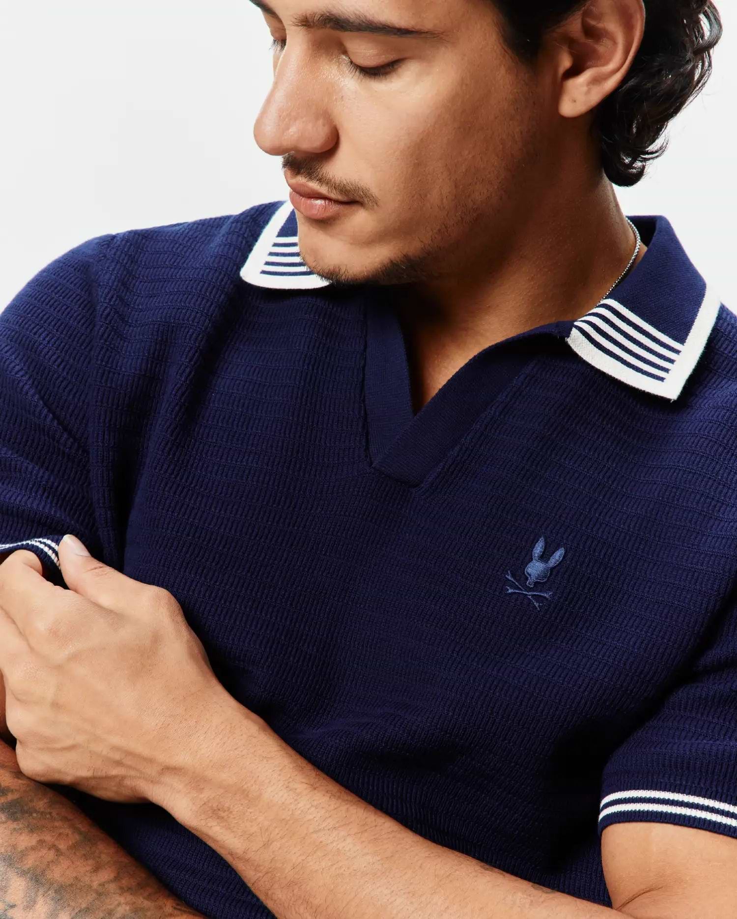Mens Elowen Knit Polo, navy blue textured knit polo with white striped collar and cuffs, featuring a small embroidered Psycho Bunny skull logo on the left chest.