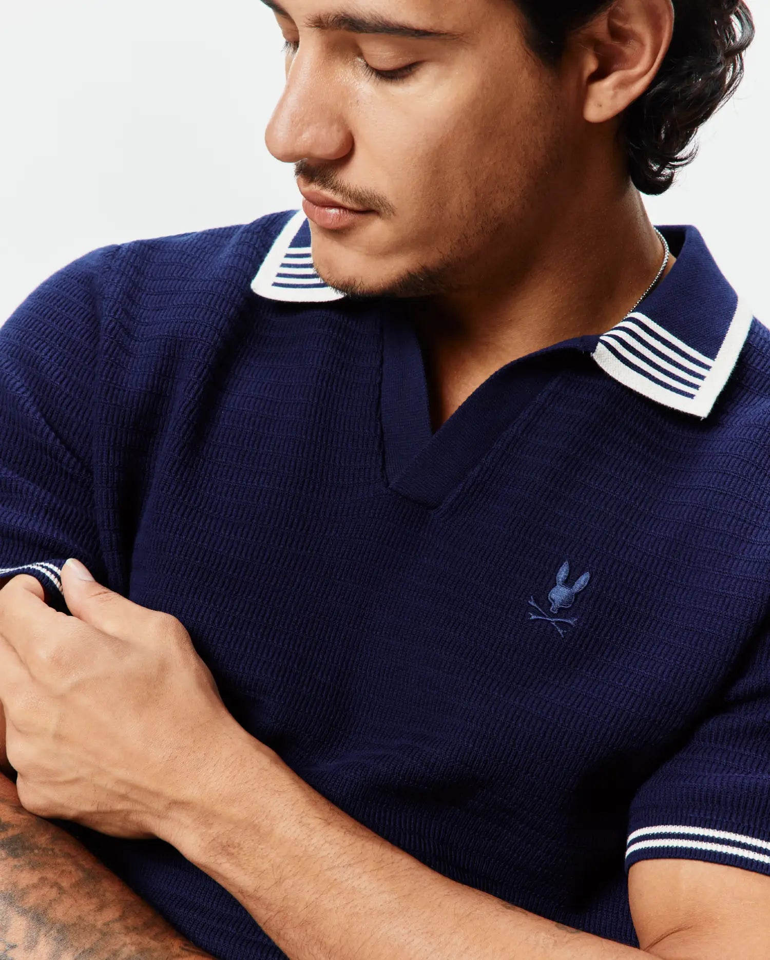 Mens Elowen Knit Polo, navy blue textured knit polo with white striped collar and cuffs, featuring a small embroidered Psycho Bunny skull logo on the left chest.