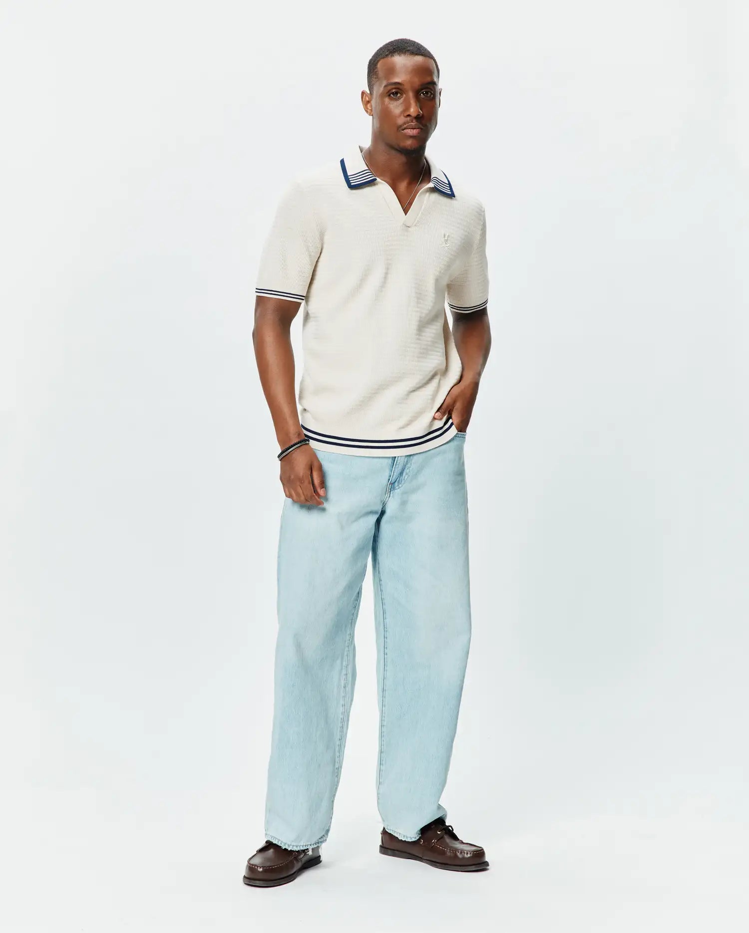 Mens Elowen Knit Polo in cream, featuring a ribbed texture, V-neck, and navy blue stripe accents on the collar, cuffs, and hem, with a small embroidered Psycho Bunny logo on the chest.