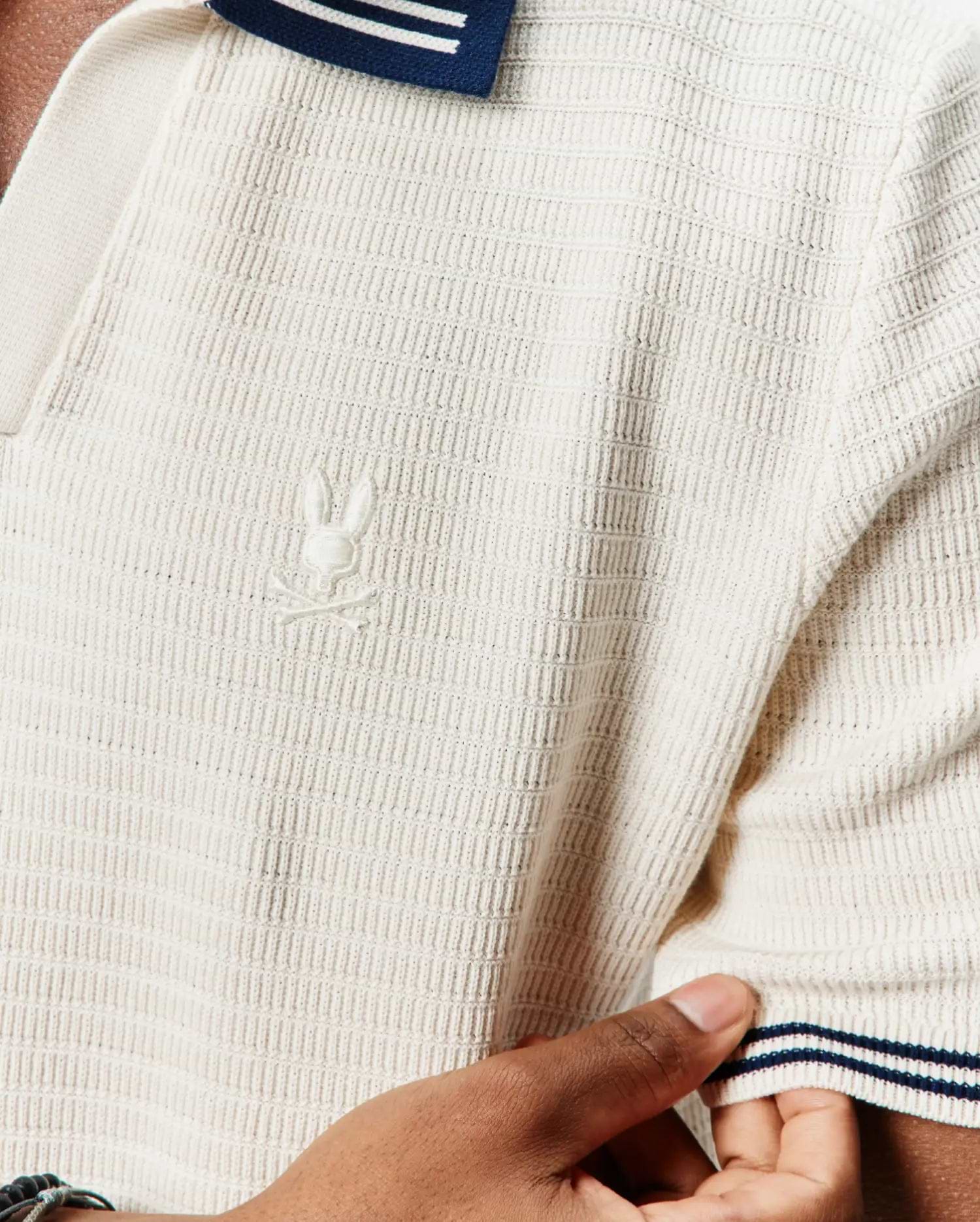 Mens Elowen Knit Polo in cream, featuring a textured knit fabric, classic fit, navy and white striped collar and sleeve cuffs, and a small embroidered Psycho Bunny skull logo on the left chest.