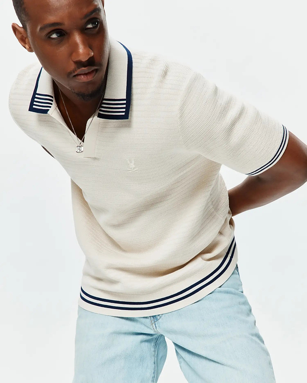 Mens Elowen Knit Polo in cream, featuring a ribbed knit texture, navy and white striped collar, cuffs, and hem, with a small embroidered Psycho Bunny skull logo on the left chest.
