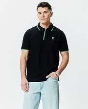 Mens Faye Pique Polo, a classic fit black pique polo shirt with a small embroidered white Psycho Bunny skull logo on the left chest, featuring green and white tipping on the collar and sleeve cuffs.
