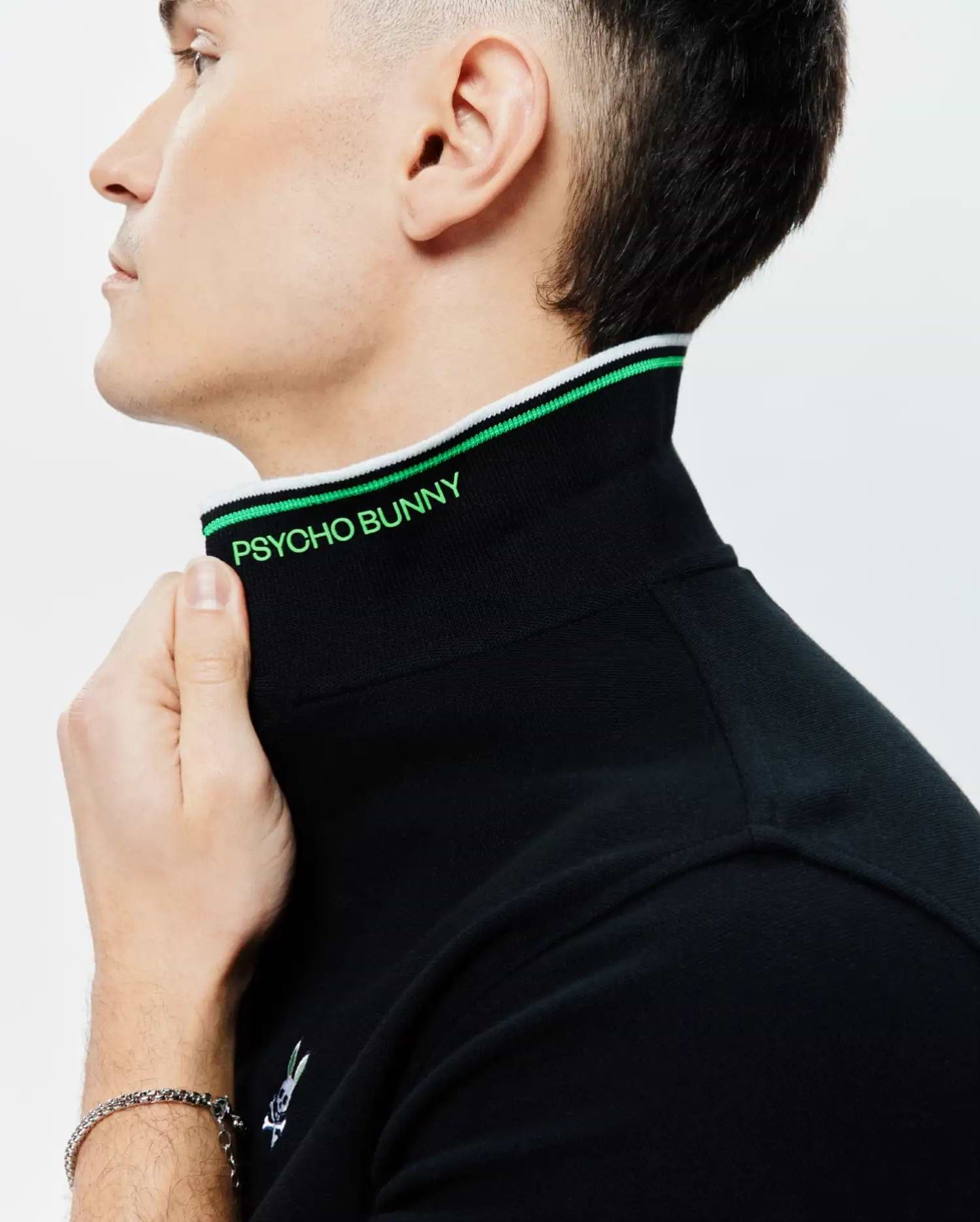 Mens Faye Pique Polo, black with a classic fit, features a ribbed collar accented with green and white stripes, bright green PSYCHO BUNNY text on the inner collar, and a small embroidered bunny skull logo.