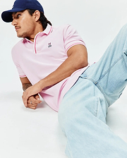 Mens Faye Pique Polo in light pink, a classic fit with textured pique fabric, featuring a small embroidered Psycho Bunny skull logo on the left chest and vibrant red striped trim on the collar and sleeve cuffs.