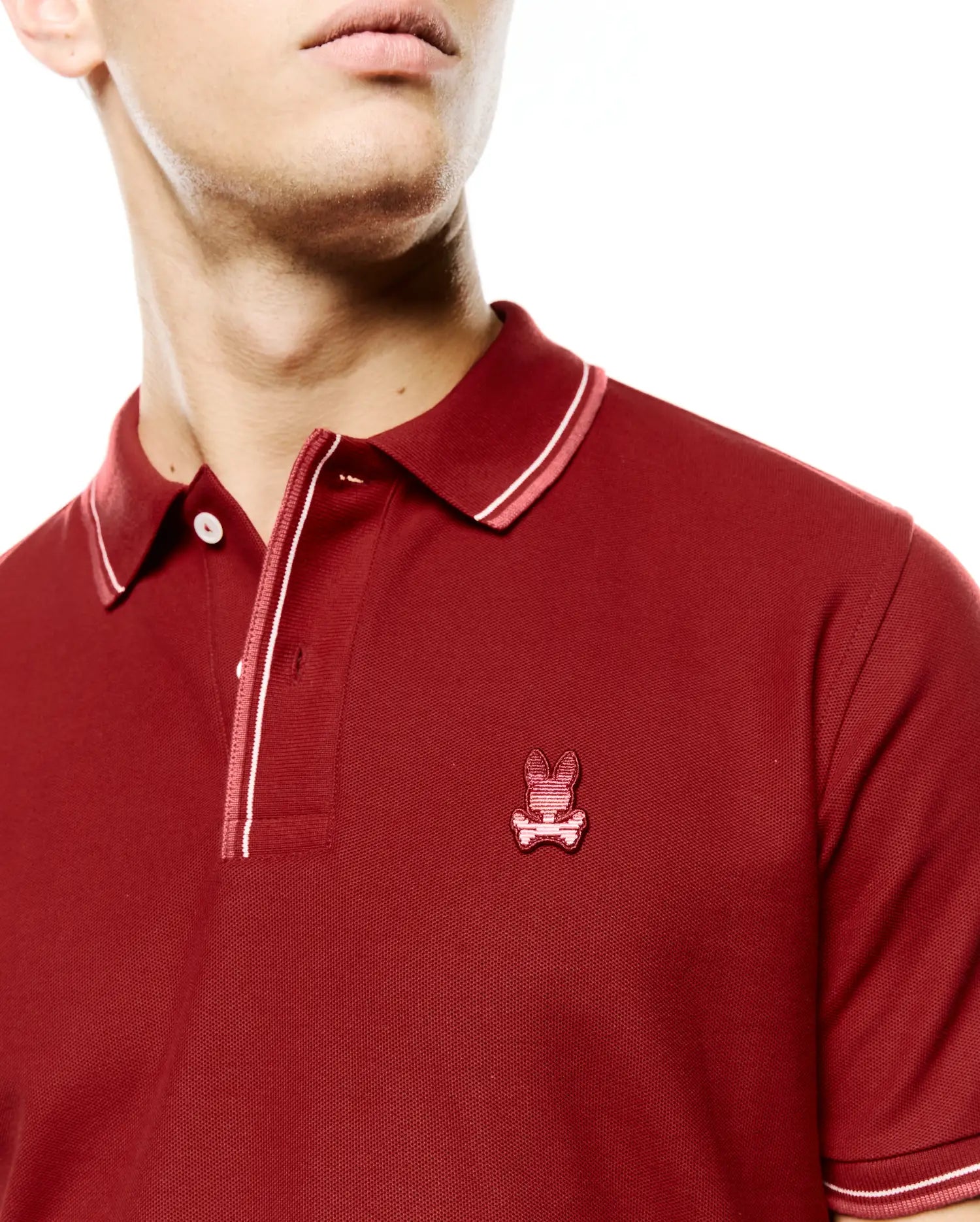 Men's Slade Pique Polo, a classic fit deep red pique polo featuring a small embroidered Psycho Bunny skull logo on the left chest and white with pink striped accents on the collar, placket, and cuffs. (2)