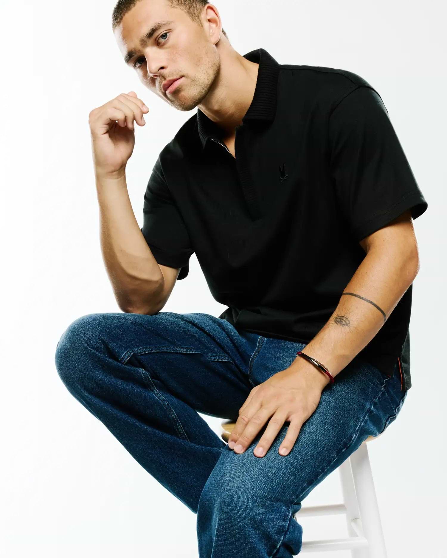 Men's Nolen Oversized Jersey Polo, a black oversized jersey polo with a ribbed collar, quarter-zip placket, small embroidered Psycho Bunny skull logo on the left chest, and subtle red side slit accents. (3)