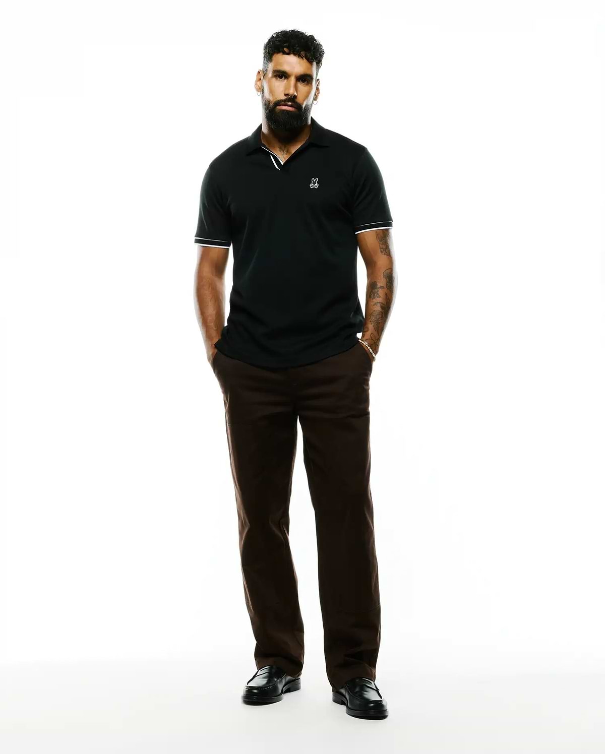 Mens Nasser Pima Johnny Polo, a black pique polo with a classic fit, featuring a small embroidered white Psycho Bunny skull logo on the left chest and white striped trim on the collar and cuffs.