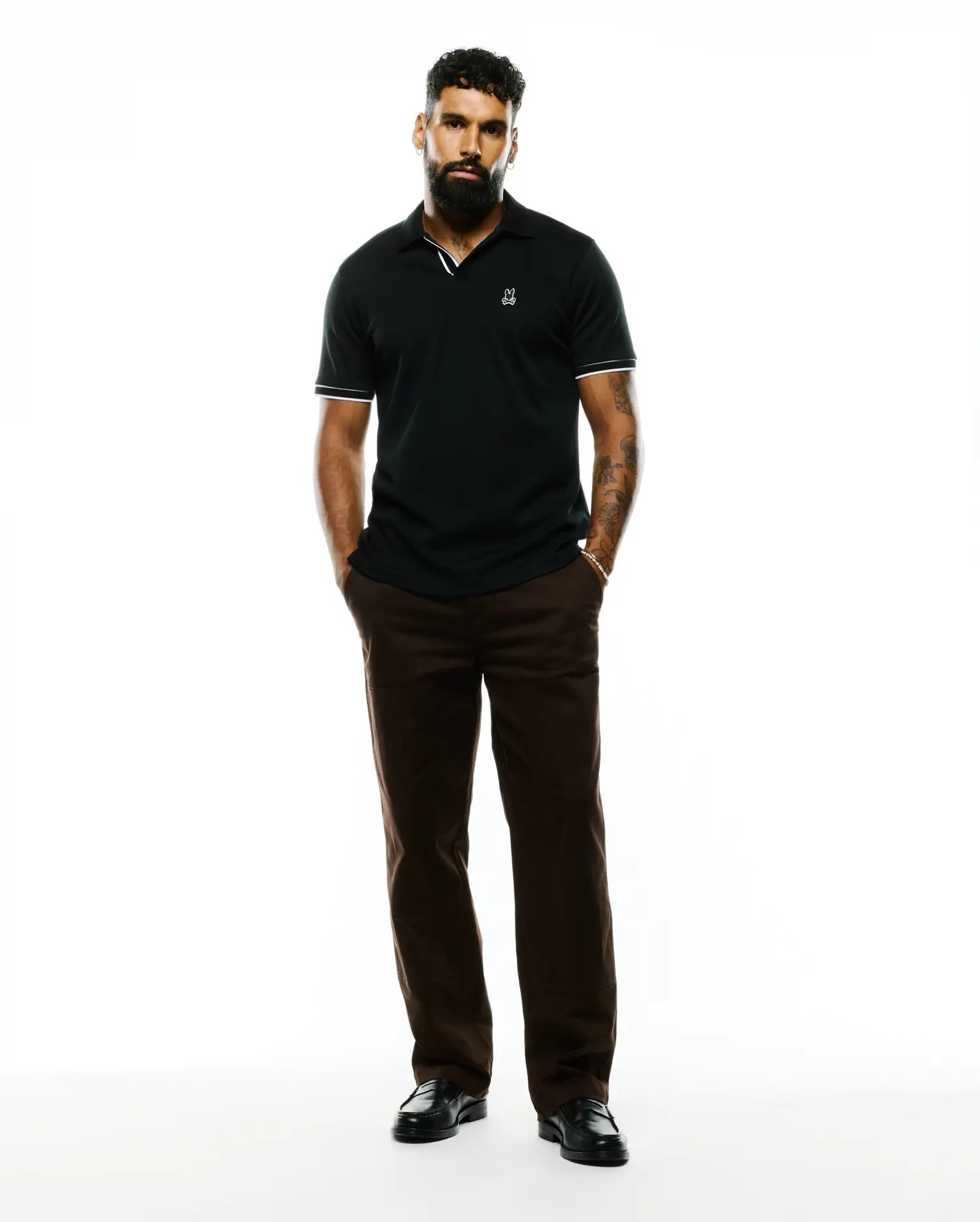 Mens Nasser Pima Johnny Polo, a black pique polo with a classic fit, featuring a small embroidered white Psycho Bunny skull logo on the left chest and white striped trim on the collar and cuffs.