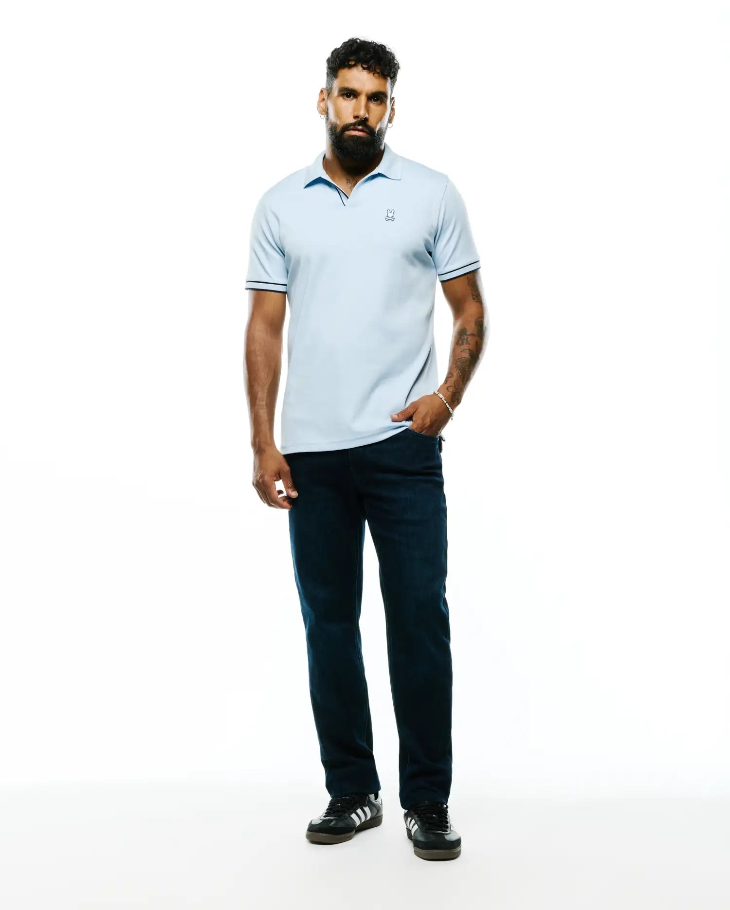 Mens Nasser Pima Johnny Polo, a light blue classic fit pique polo with an embroidered Psycho Bunny skull logo on the left chest and navy tipping on the collar and sleeve cuffs.